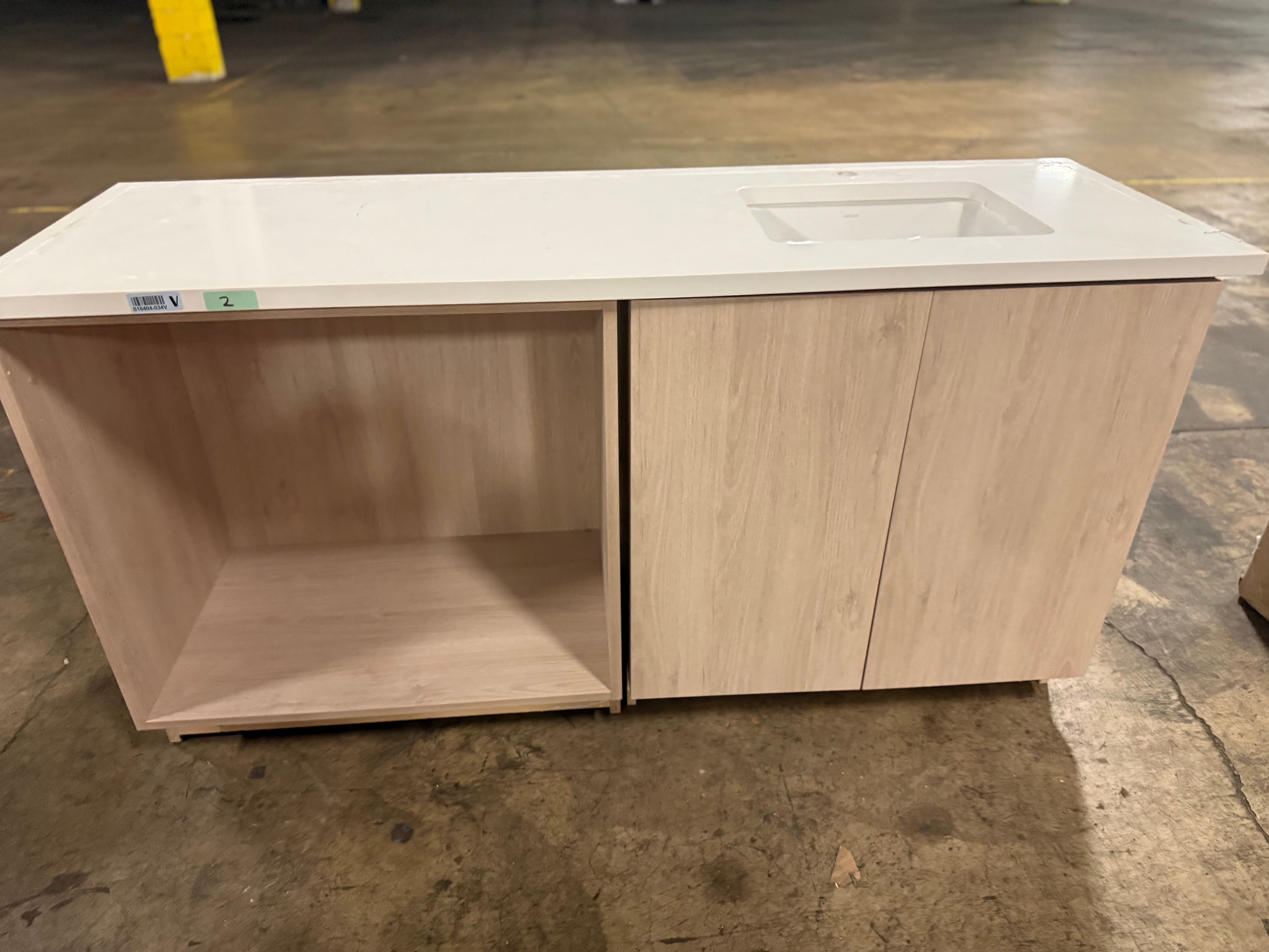 A light wood kitchen island cabinet with a white countertop and an integrated sink, photographed in a warehouse setting on a concrete floor.
