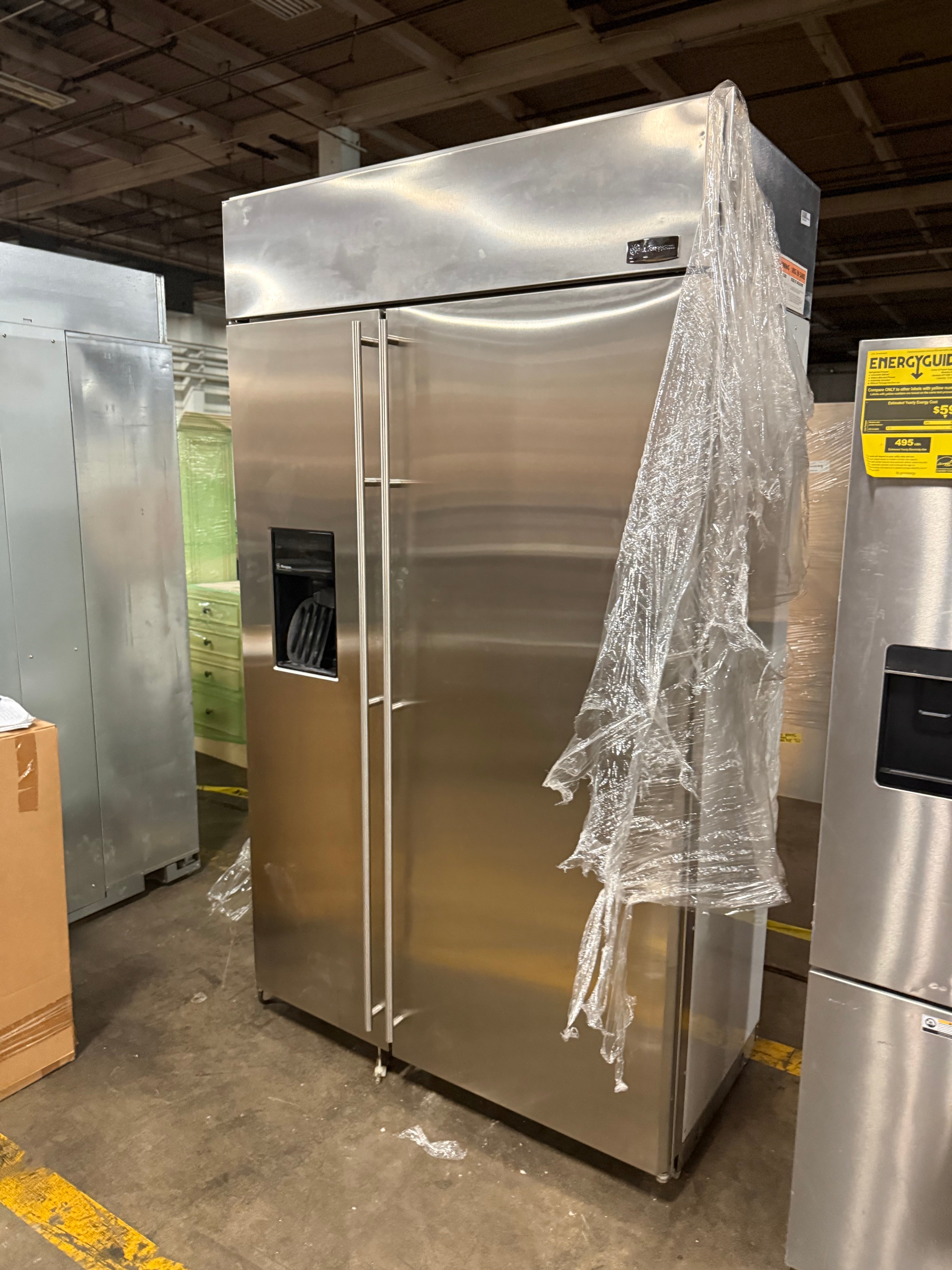 A large stainless steel commercial refrigerator with two doors, a side water dispenser, and a sheet of clear bubble wrap hanging from the right side, photographed in a warehouse setting.