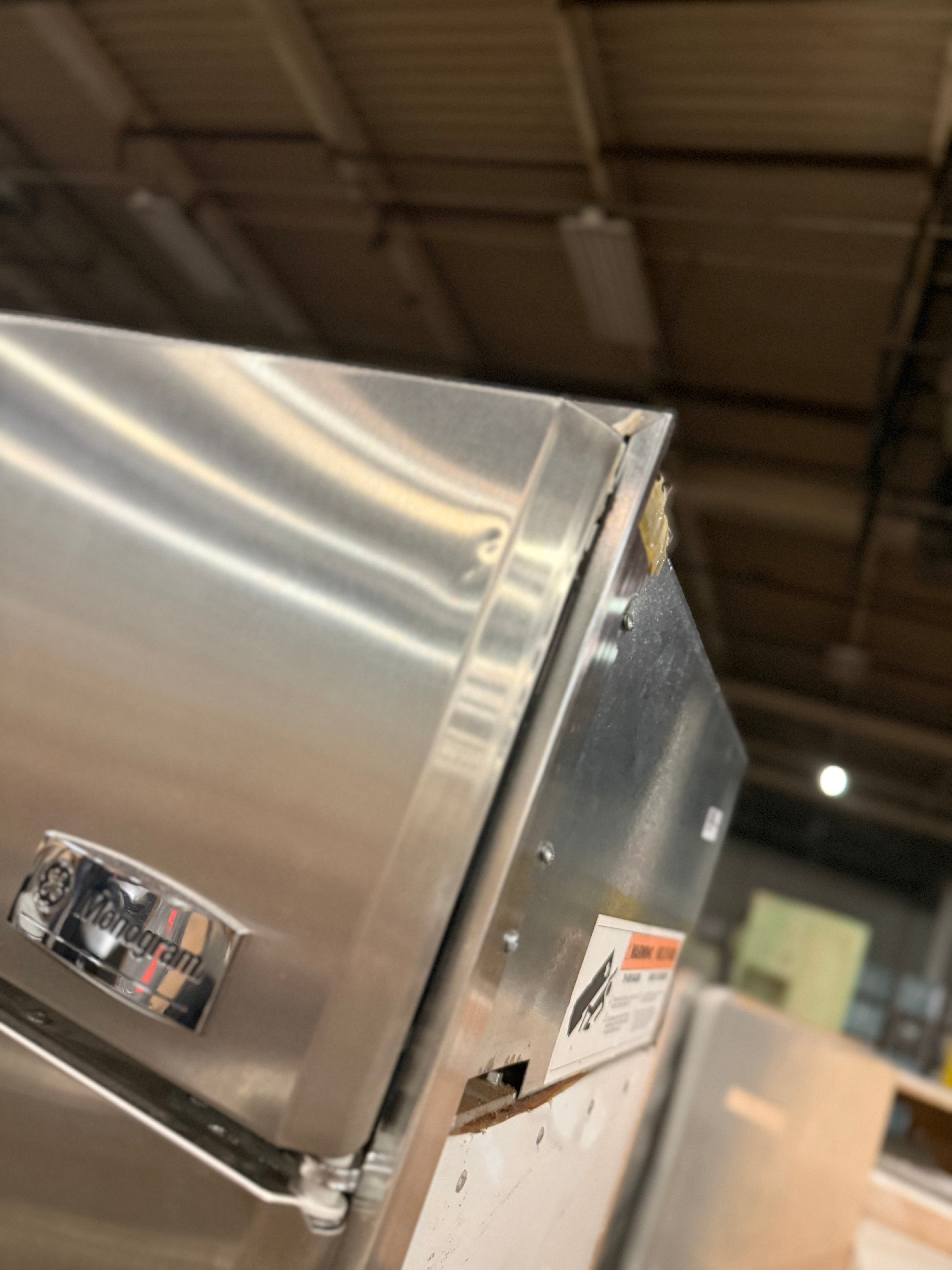 A close-up, angled view of a stainless steel commercial refrigerator or freezer, showing the side panel, hinge, and a small 'Kemper' brand badge. The background is a blurry warehouse or showroom environment with ceiling lights.