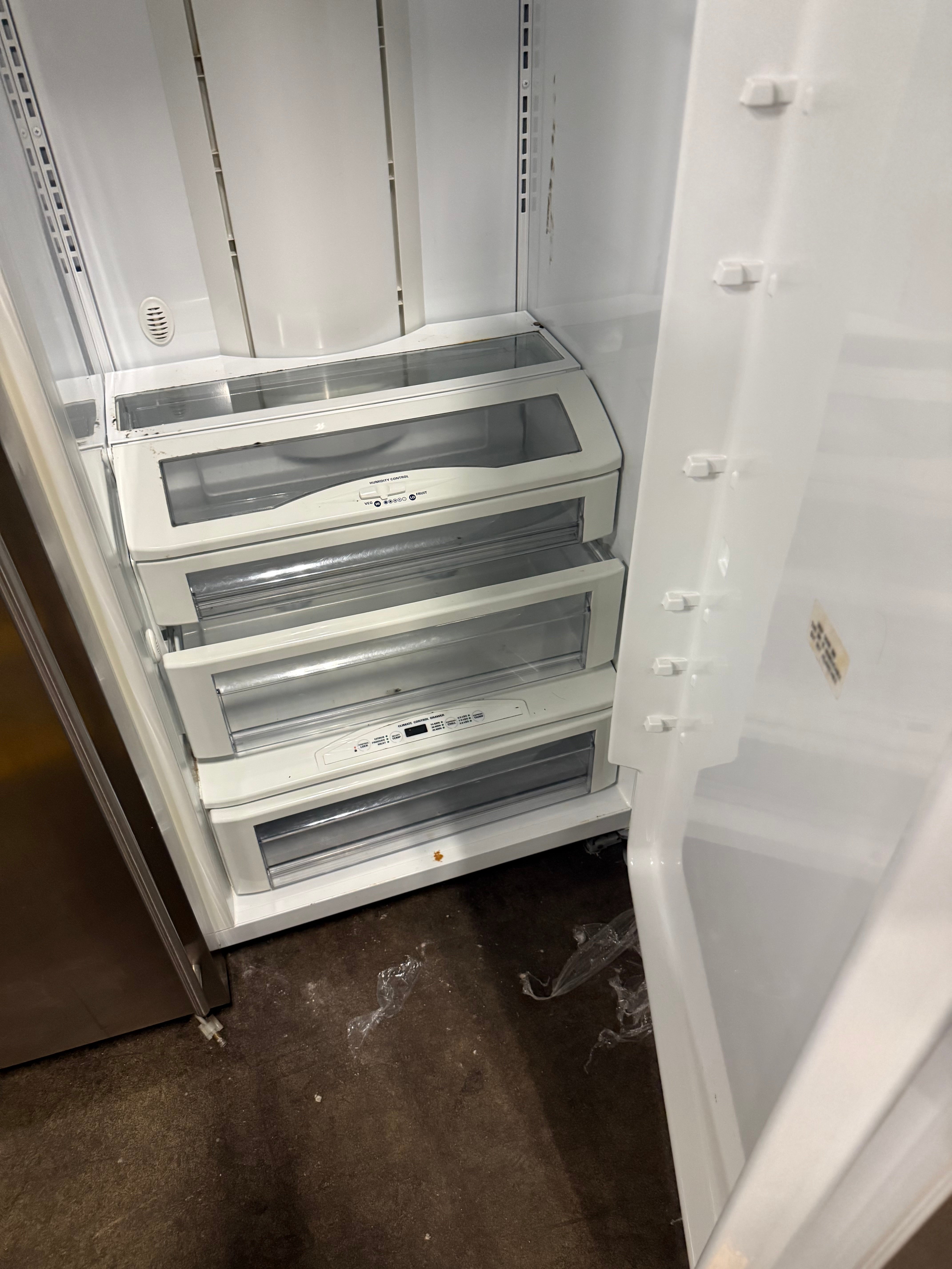 An open white refrigerator showing the freezer drawer compartment with three transparent-front crisper drawers. The interior is empty, and the floor is a dark brown surface.