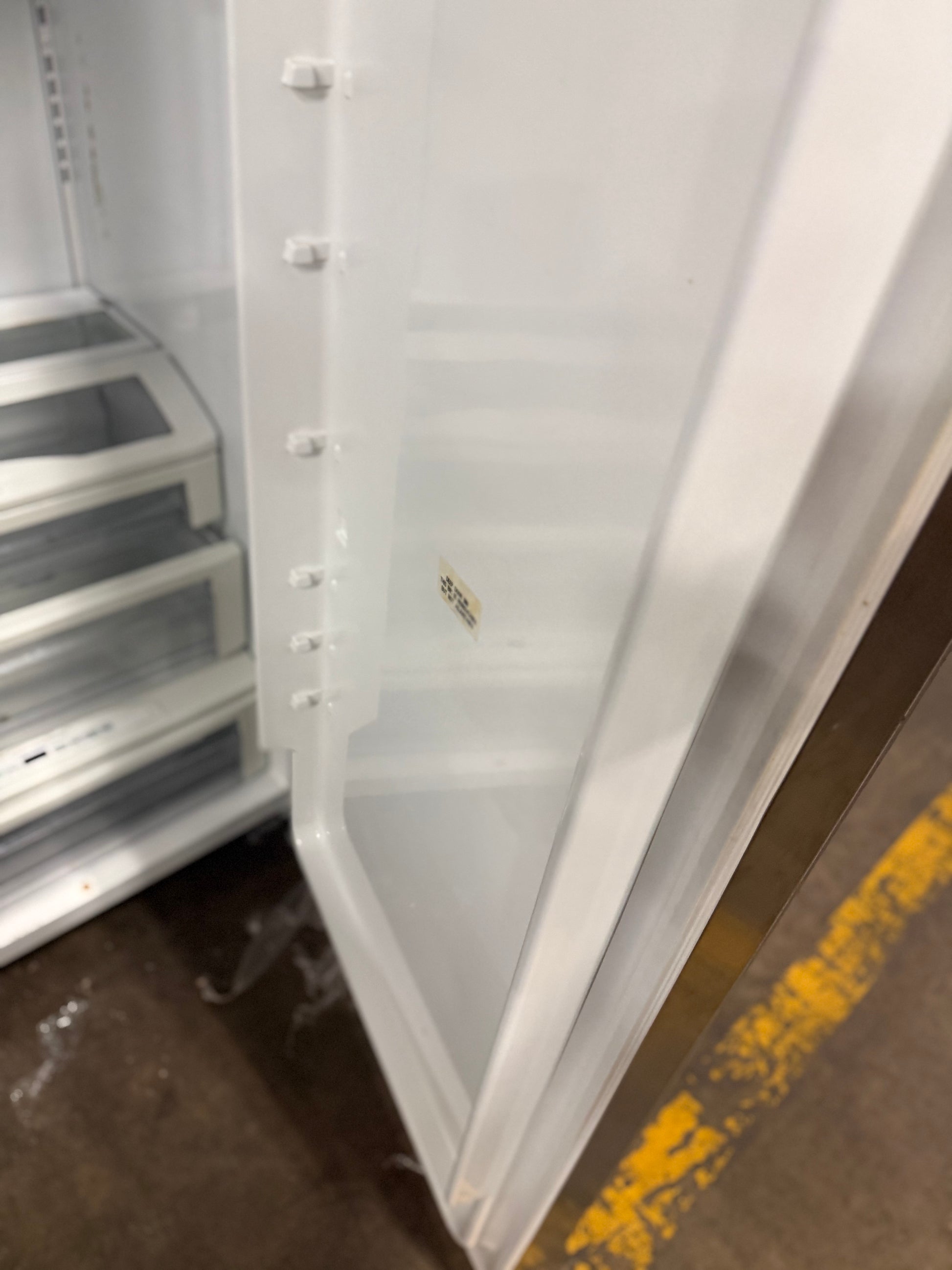 A close-up view of an empty white refrigerator door with its drawer compartment open, showing the interior shelves and a small sticker on the inner door, set against a warehouse floor with a yellow stripe.