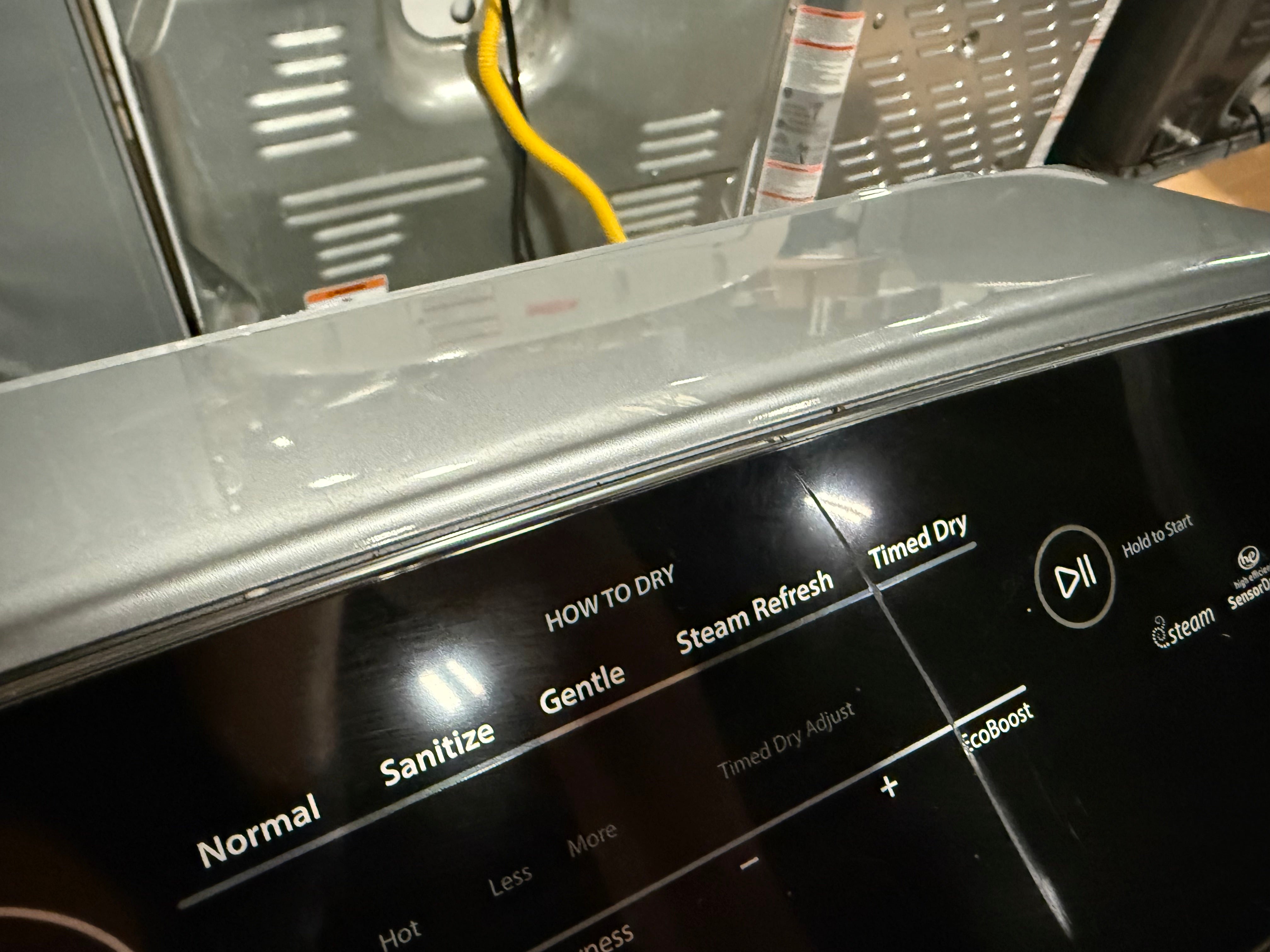 Close-up of a black and silver dryer control panel with touch buttons and text options like Normal, Sanitize, Gentle, Steam Refresh, and Timed Dry. A yellow utility cord and metal appliance interior are visible in the background.