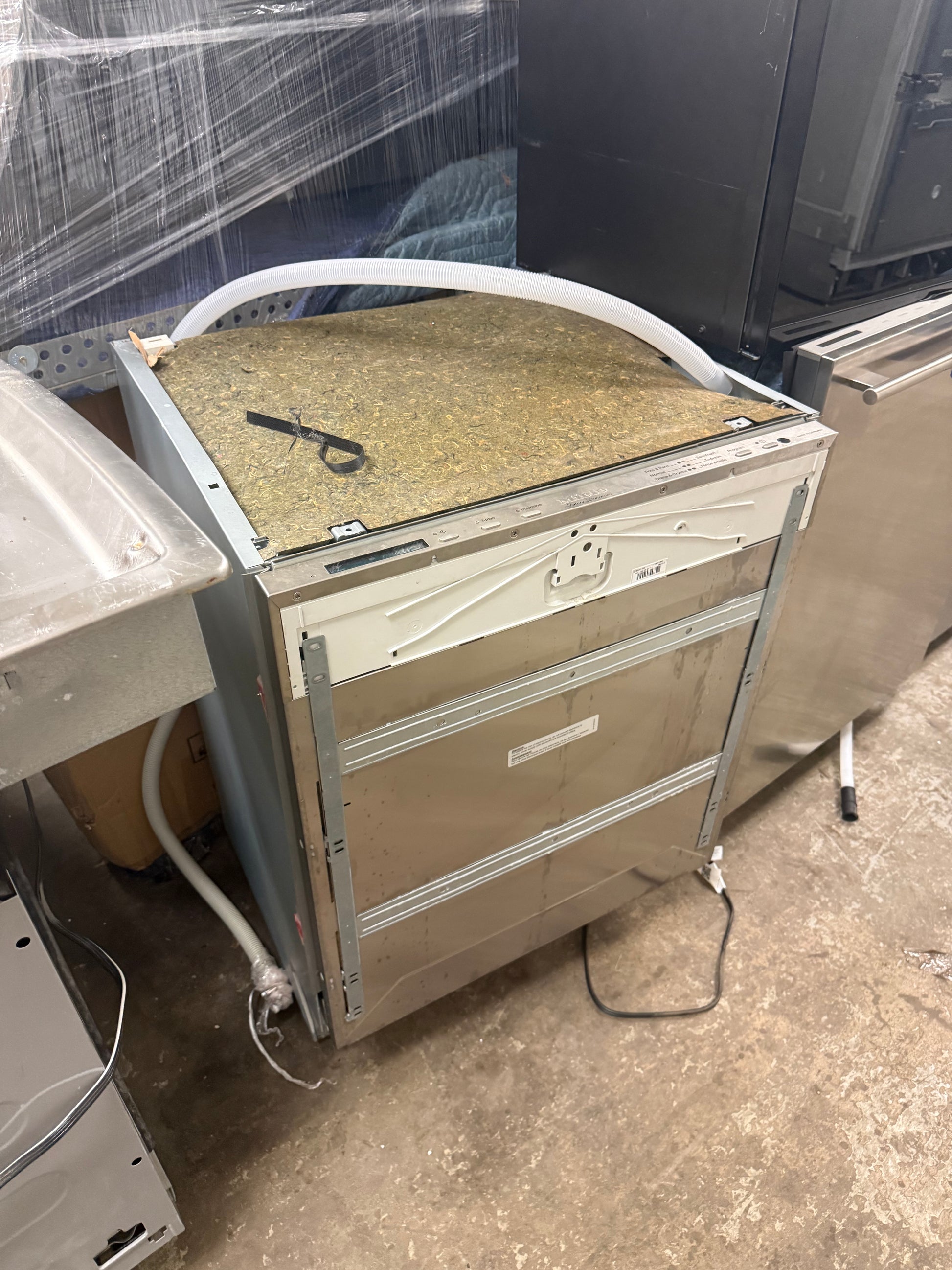 A stainless steel undercounter dishwasher shown from the back, with its top panel removed to reveal the interior and a white supply hose connected. It is photographed in a warehouse or appliance repair shop setting.