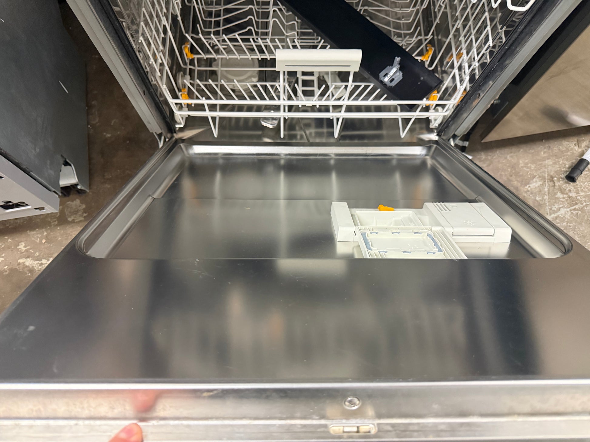 An open stainless steel dishwasher with the door swung open, showing the interior racks and a small instruction booklet on the bottom tray.