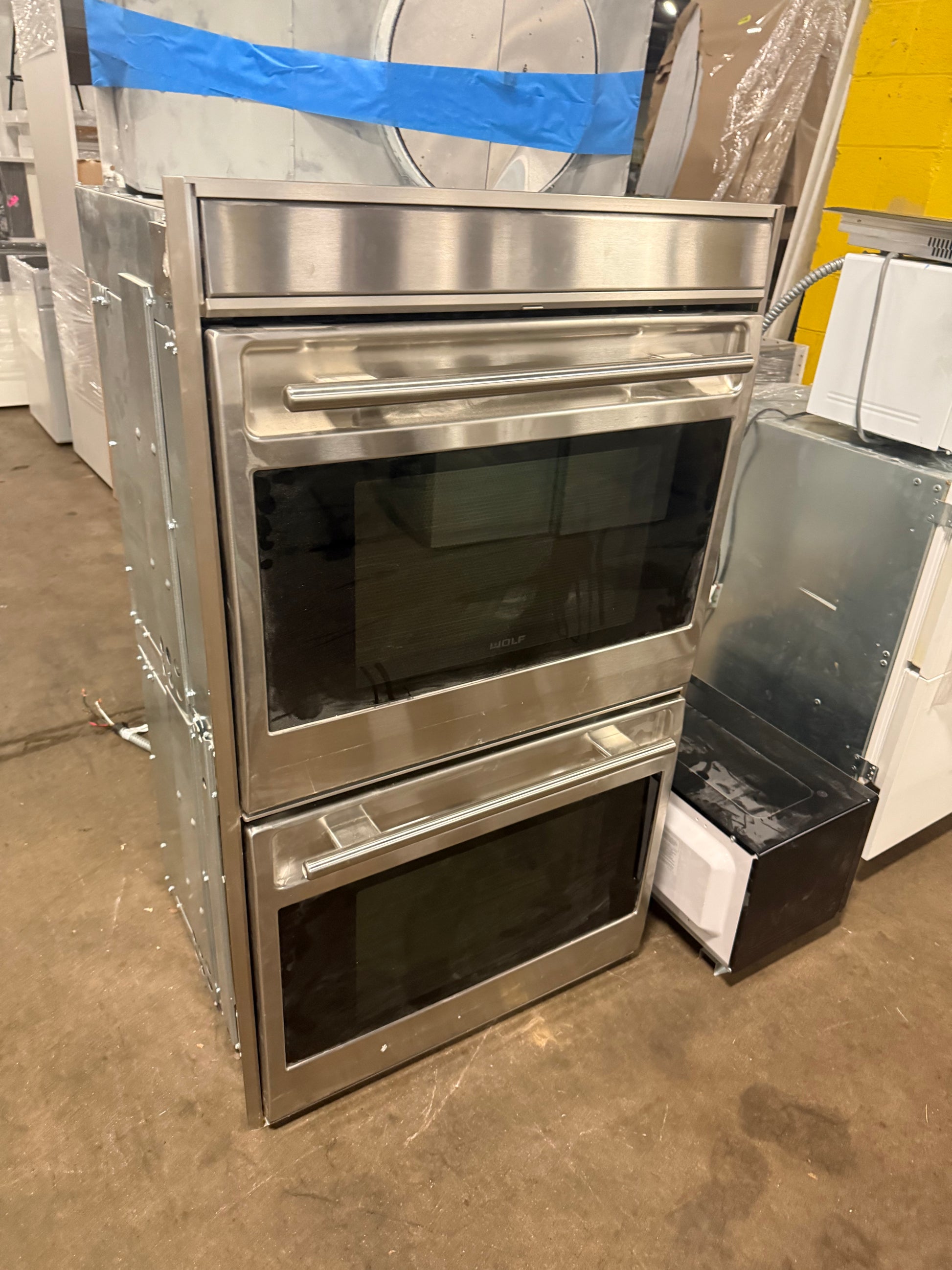 A stainless steel double wall oven with two glass-front doors and long horizontal handles, photographed in a warehouse or showroom setting with other appliances and wrapping materials visible around it.