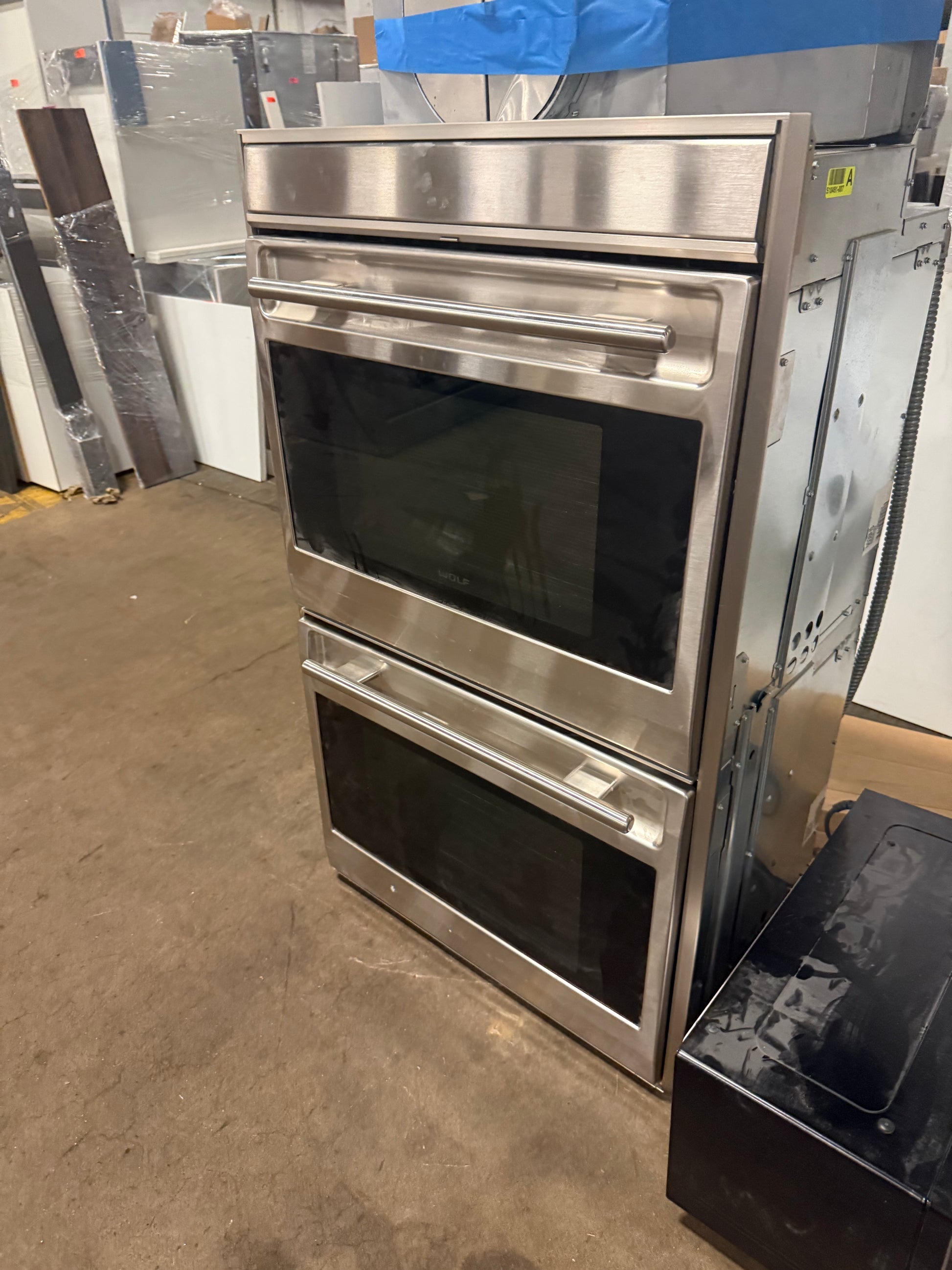 A stainless steel double wall oven with a lower drawer, photographed in a warehouse or appliance showroom setting with other appliances and boxes visible in the background.