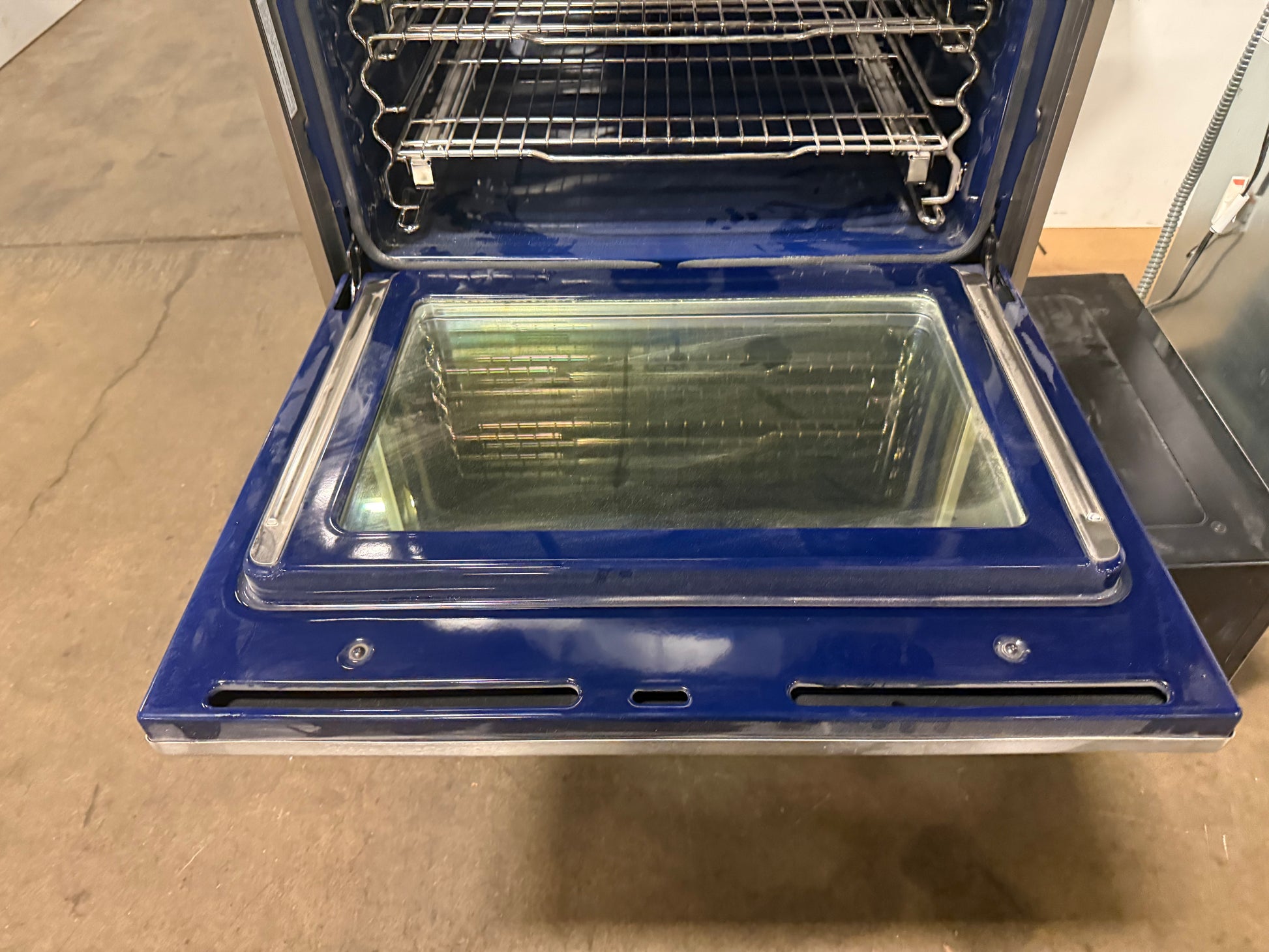 A blue electric oven with its door open, showing the interior racks and glass door window, photographed in a warehouse or workshop setting on a concrete floor.