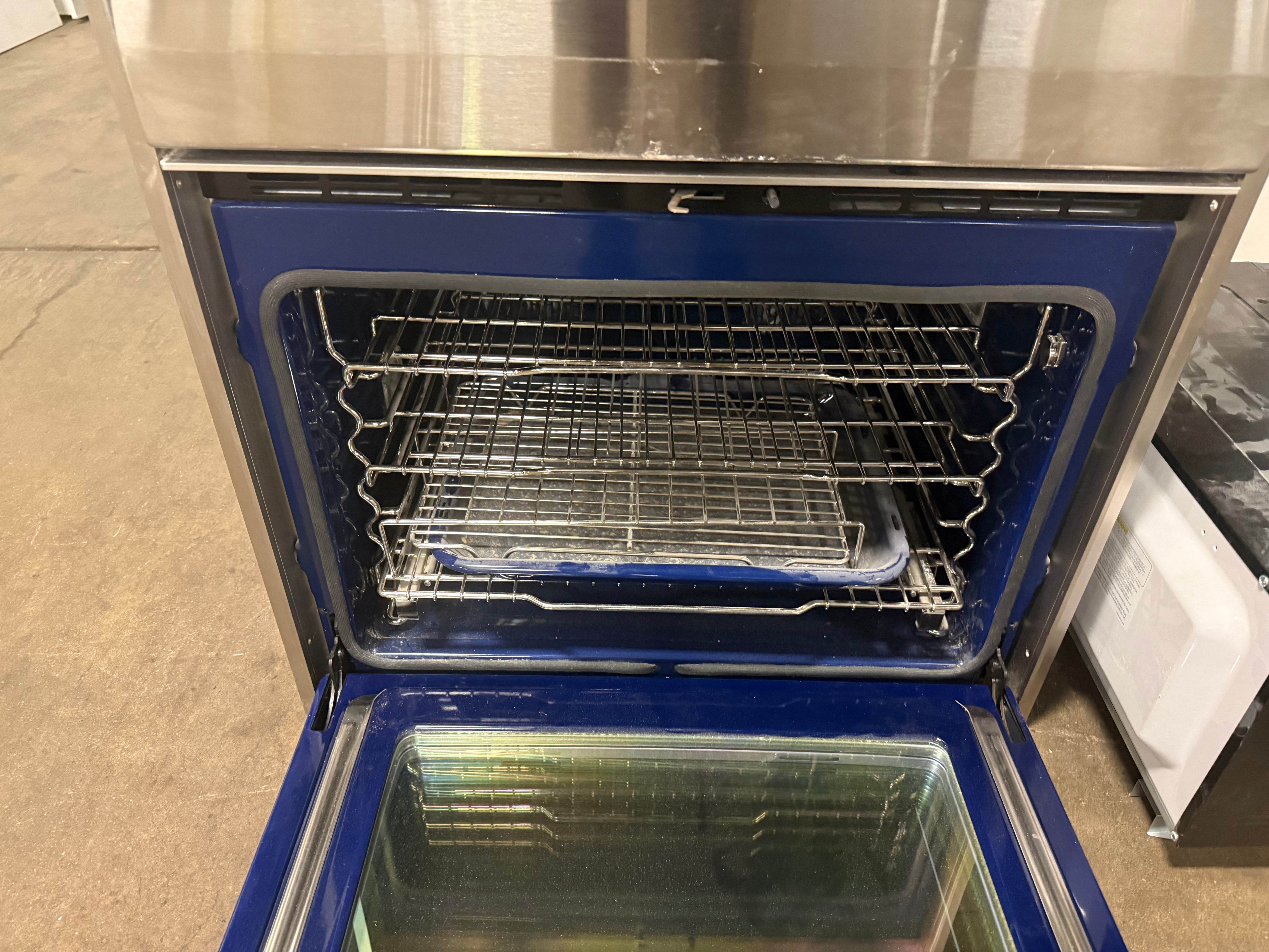 An open stainless steel and blue oven or refrigerator door showing the interior racks and glass door, photographed in a warehouse or showroom setting.
