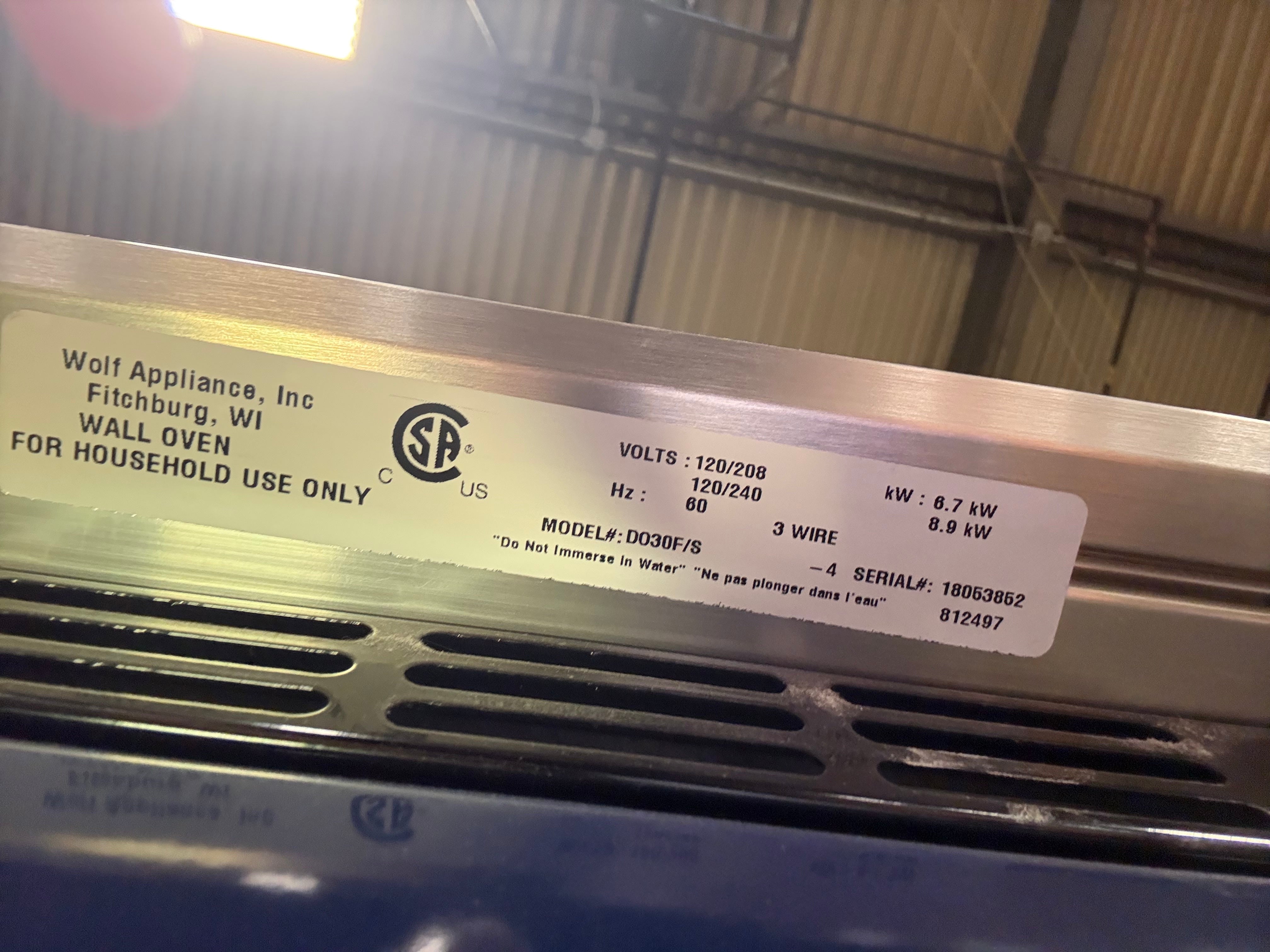 A close-up of a metal appliance label reading Wolf Appliance, Inc, with model information and a CSA mark, photographed in an industrial setting.