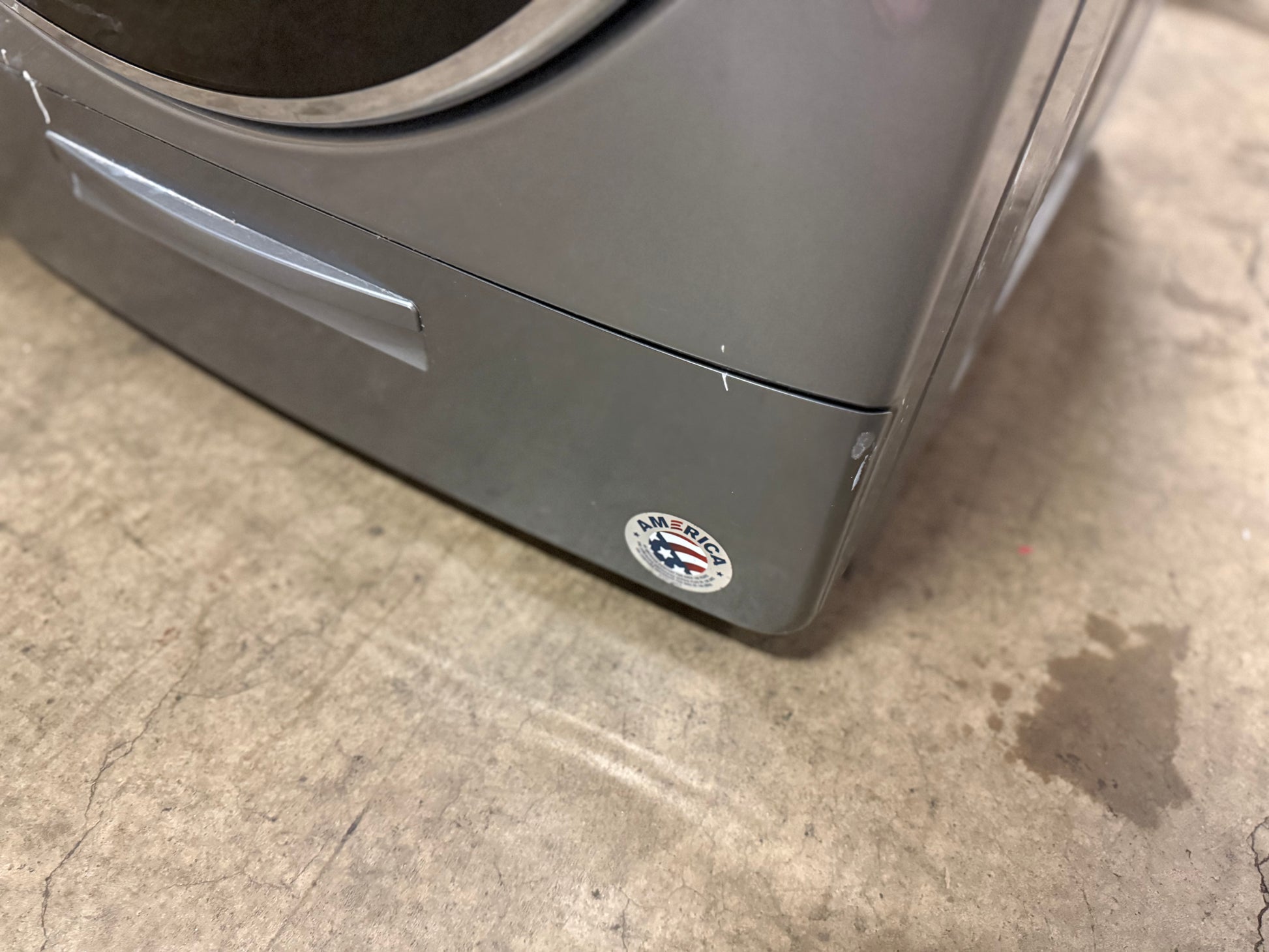Close-up of the lower corner of a gray washing machine on a concrete floor, showing a small circular sticker on the front panel.