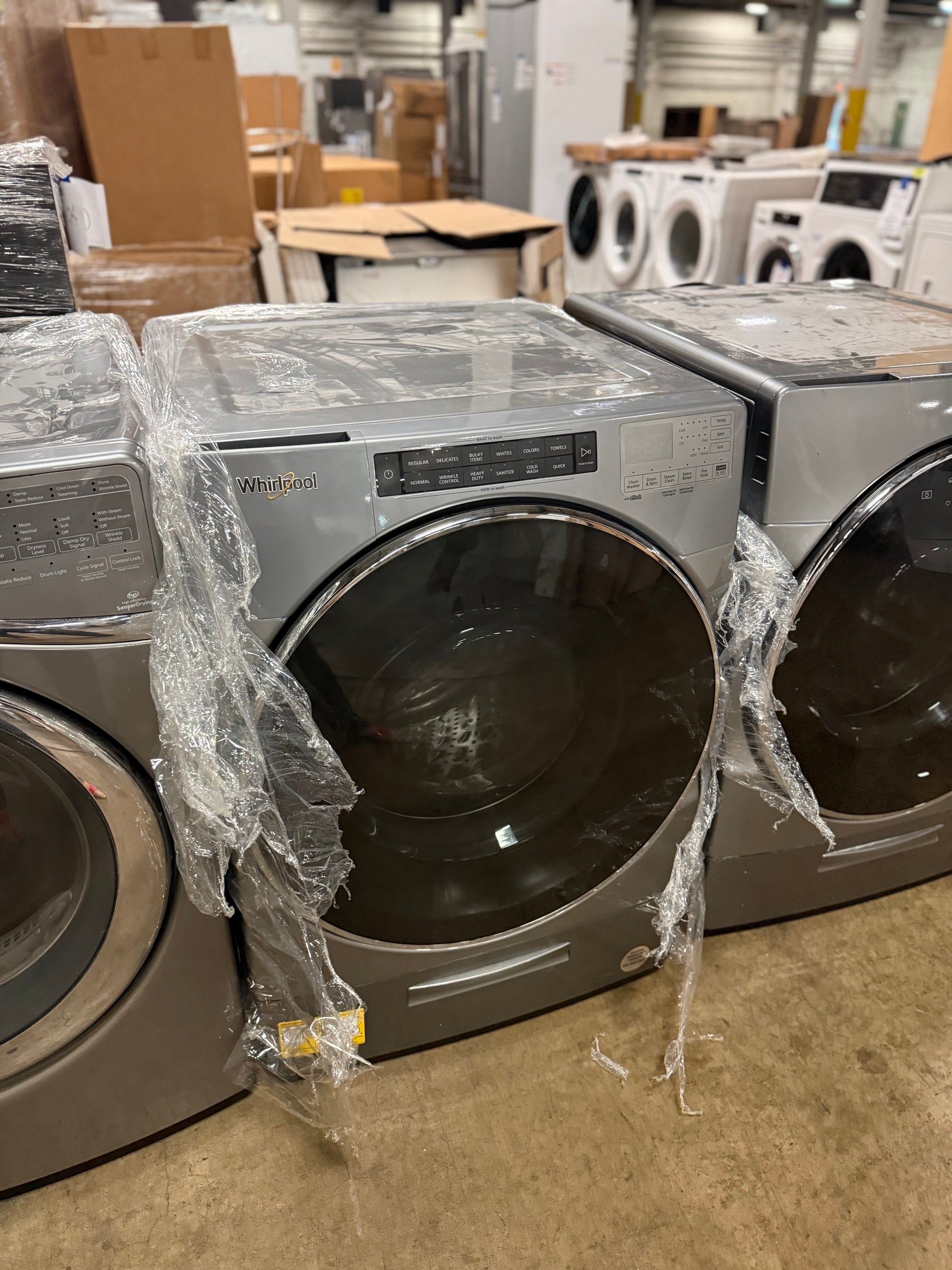 A front-load clothes dryer in a warehouse or appliance showroom setting, wrapped in protective plastic on the sides. The dryer has a metallic gray/silver finish and a large dark glass door.