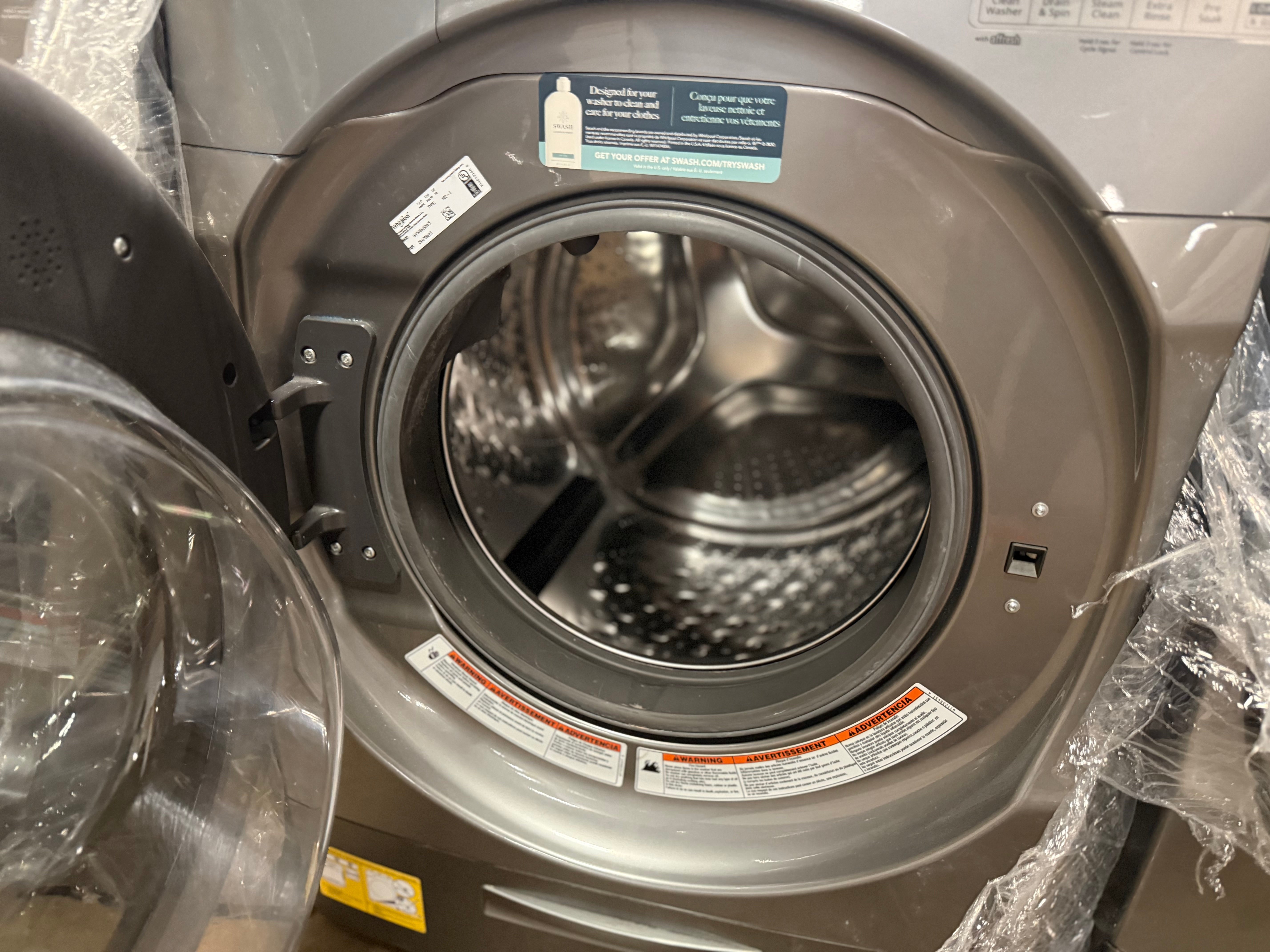 A front-loading washing machine with its door open, showing the stainless steel drum inside. The exterior has a dark gray metallic finish, and protective plastic wrapping is visible around the door and edges.