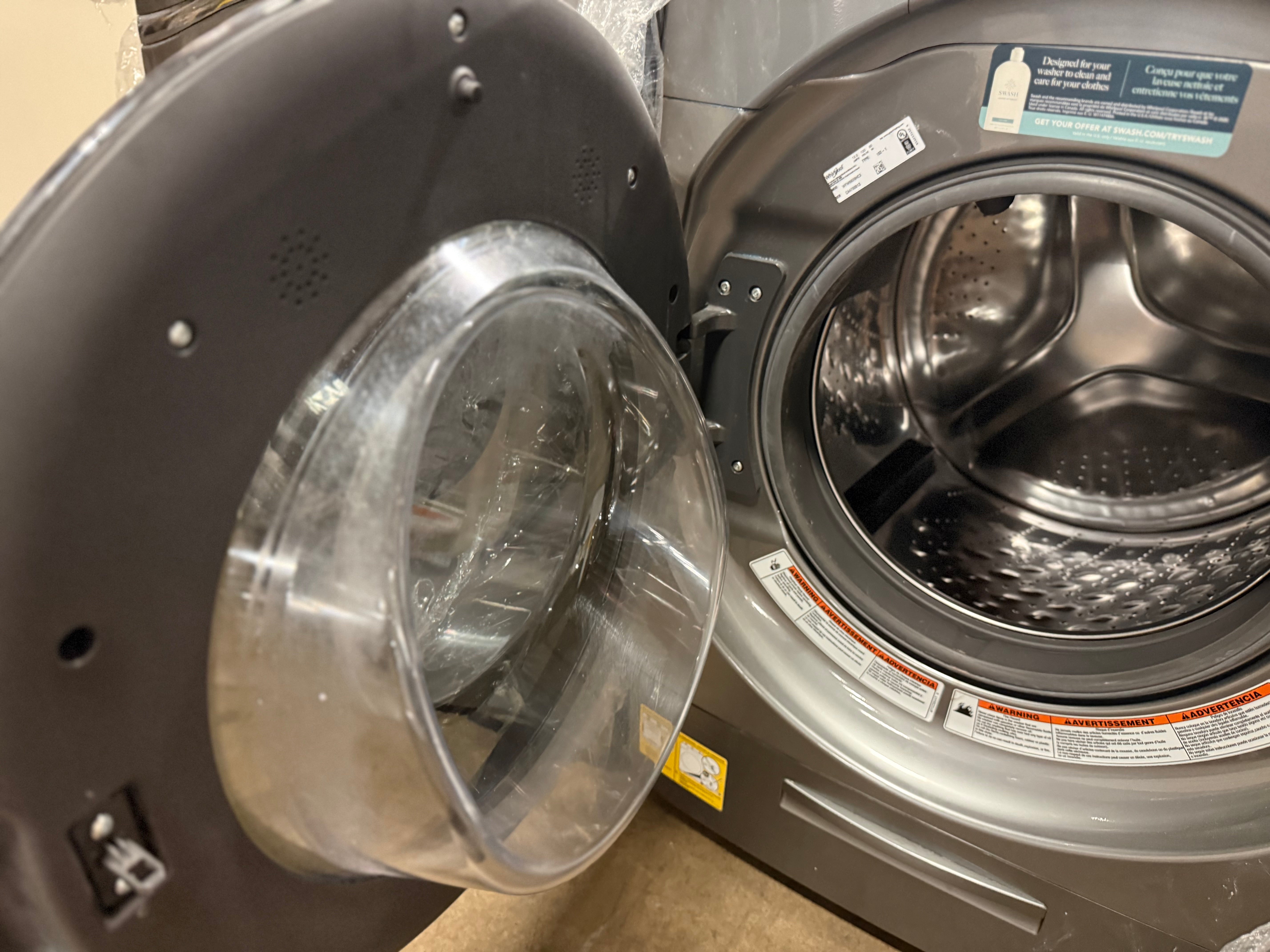 A close-up of a front-load washing machine with the door open, showing the transparent circular door seal and the stainless steel drum inside.