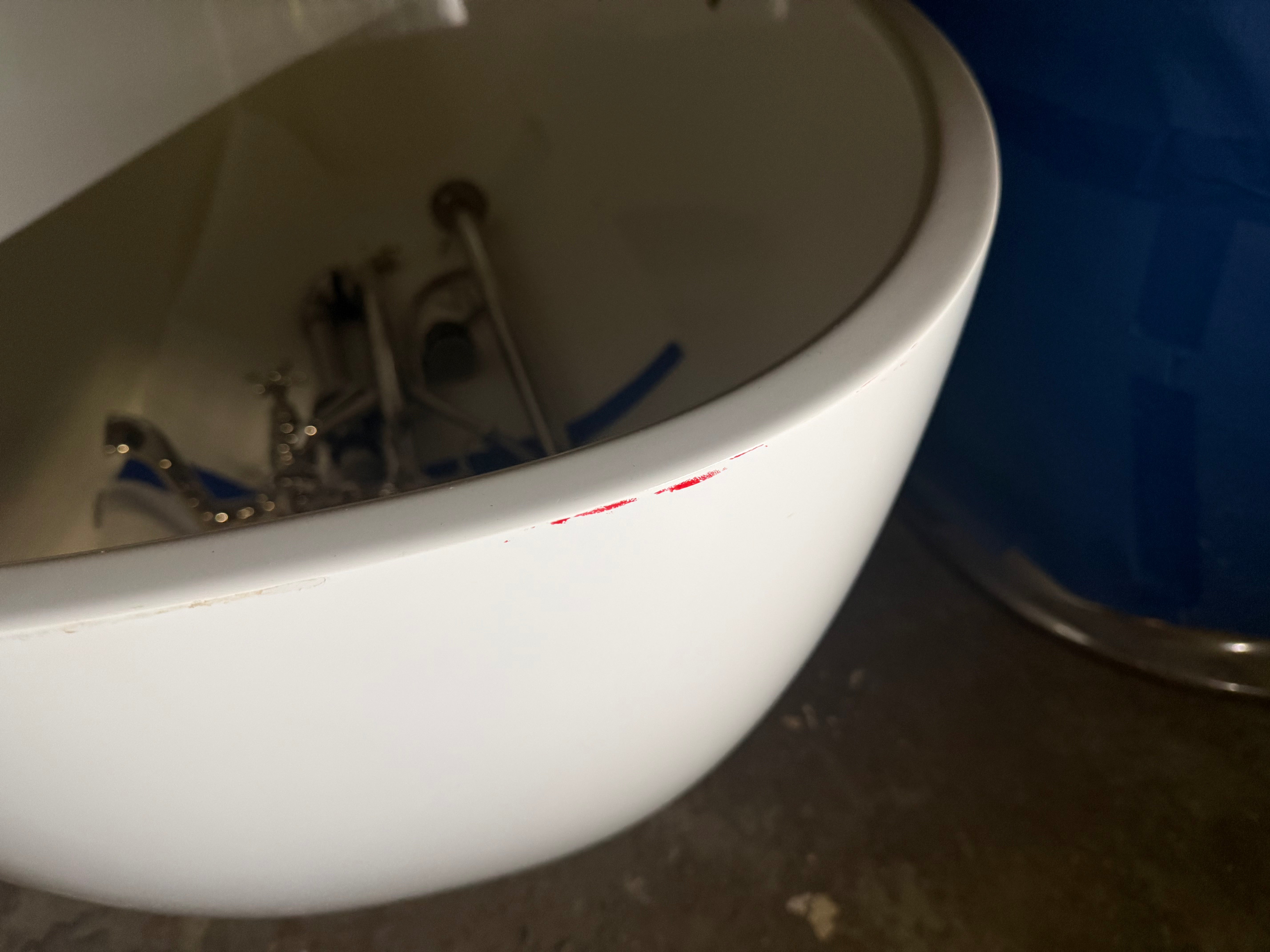 A close-up of a white ceramic wall-mounted sink basin showing the interior plumbing and a small red scratch on the rim, next to a blue barrel.