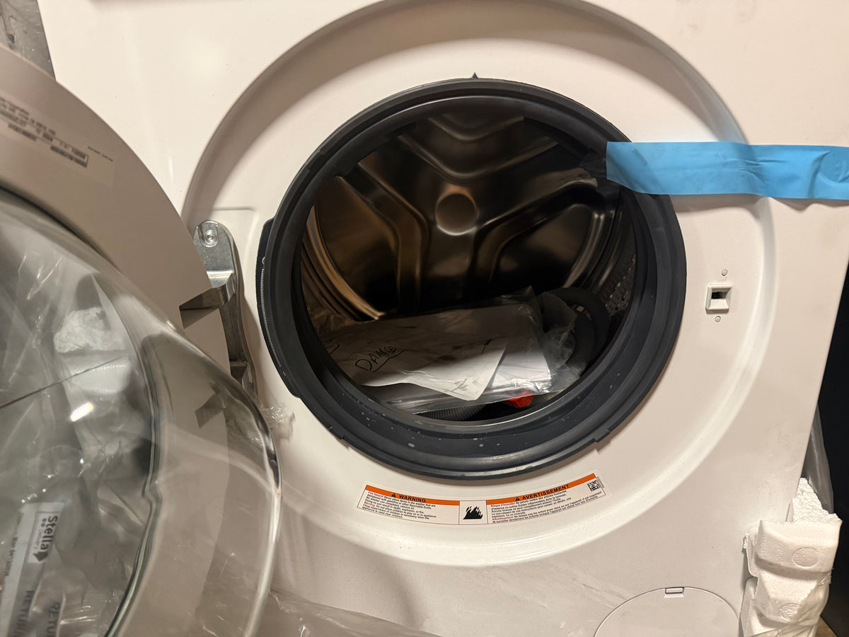 A close-up of a white front-loading washing machine with the door open, showing the dark drum interior and some laundry inside. A blue tape strip is visible along the top edge of the opening.