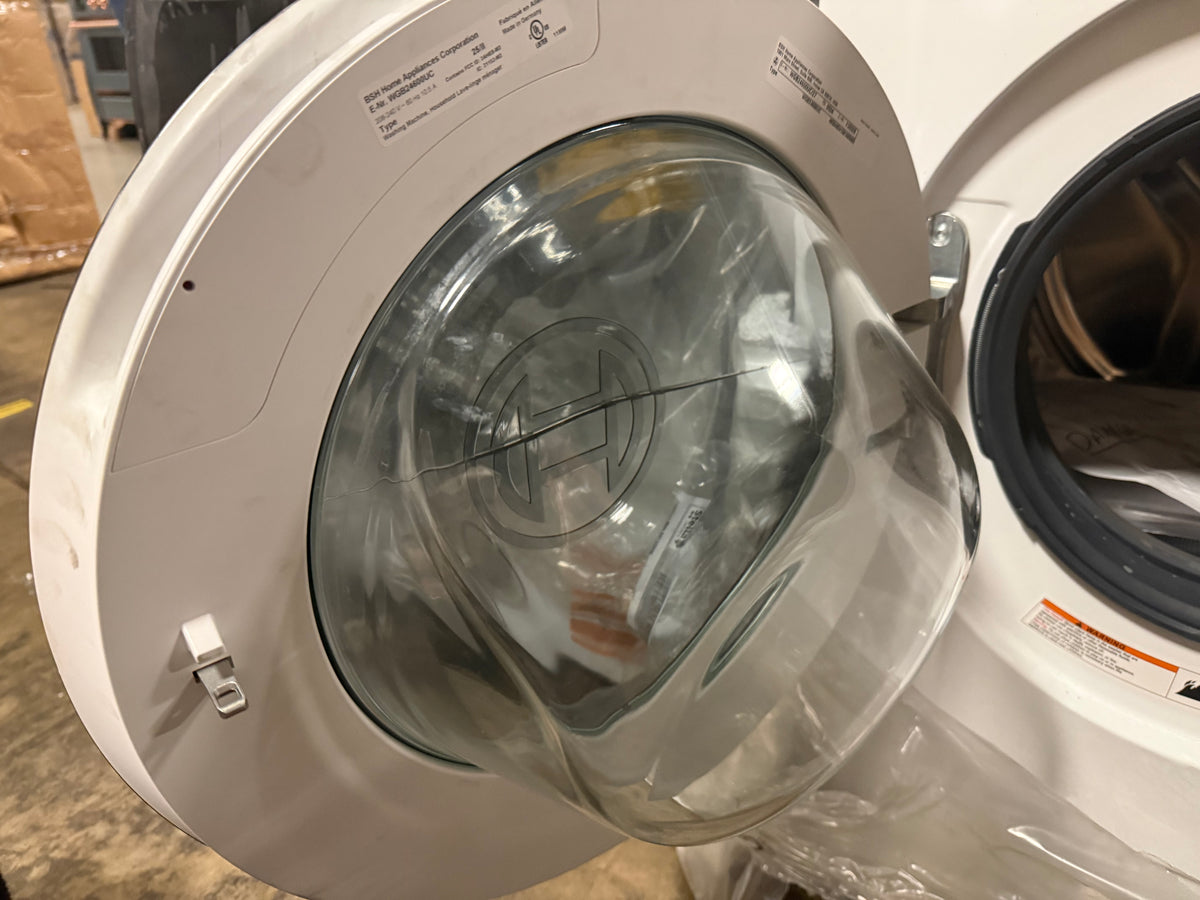 Close-up of a white front-loading washing machine with the door open, showing the clear glass door and drum interior in a warehouse-like setting.