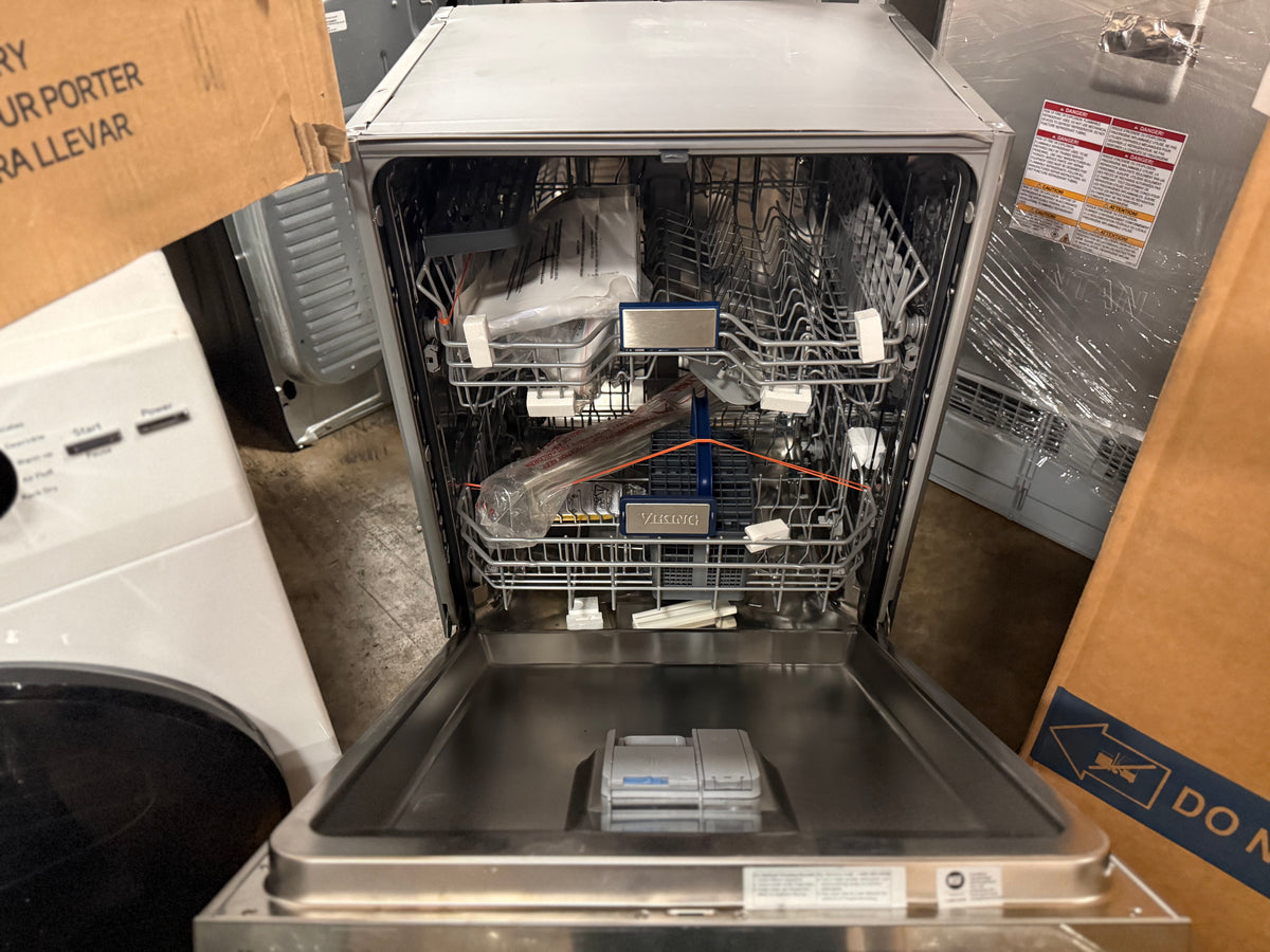 A stainless steel built-in dishwasher with its door open, showing the interior racks and detergent dispenser, photographed in a showroom or warehouse setting with other appliances and cardboard boxes nearby.