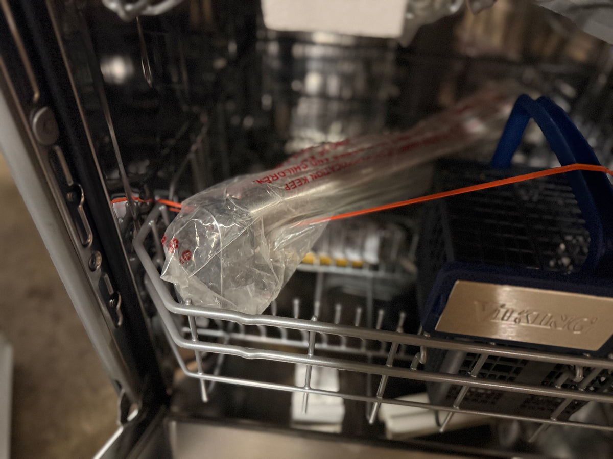 A close-up view inside an open dishwasher, showing a packaged metal utensil or tool resting on the lower rack next to a blue-handled item. The dishwasher interior is metallic and utilitarian.