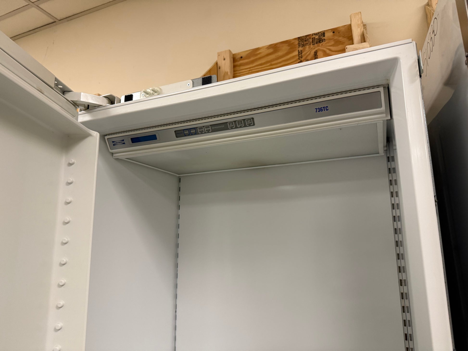 A white metal server rack cabinet with an open top door, showing a mounting bracket installed inside. The cabinet is photographed in a warehouse or workshop setting with a beige wall and wooden pallet visible in the background.