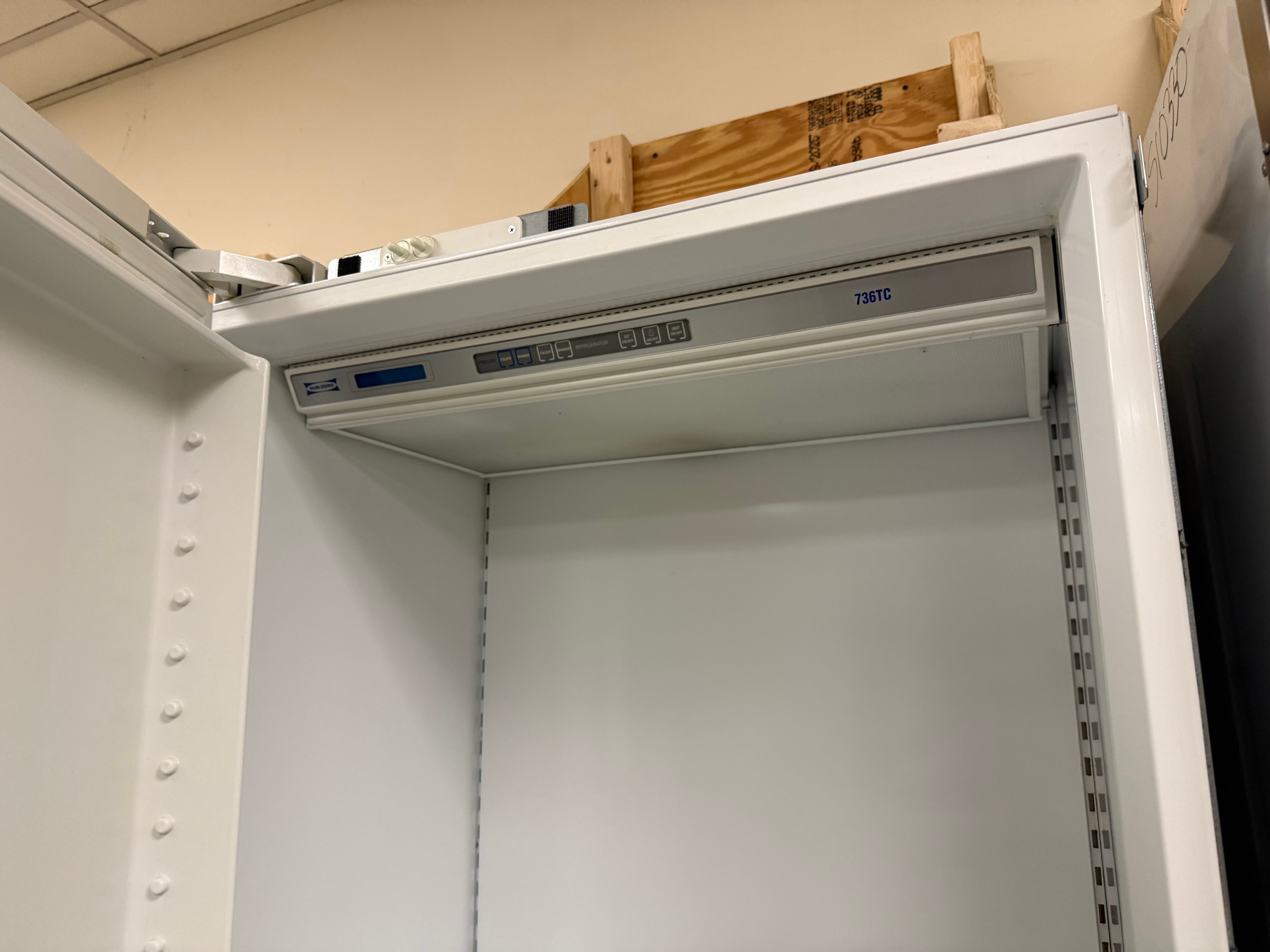 A white metal server rack cabinet with an open top door, showing a mounting bracket installed inside. The cabinet is photographed in a warehouse or workshop setting with a beige wall and wooden pallet visible in the background.
