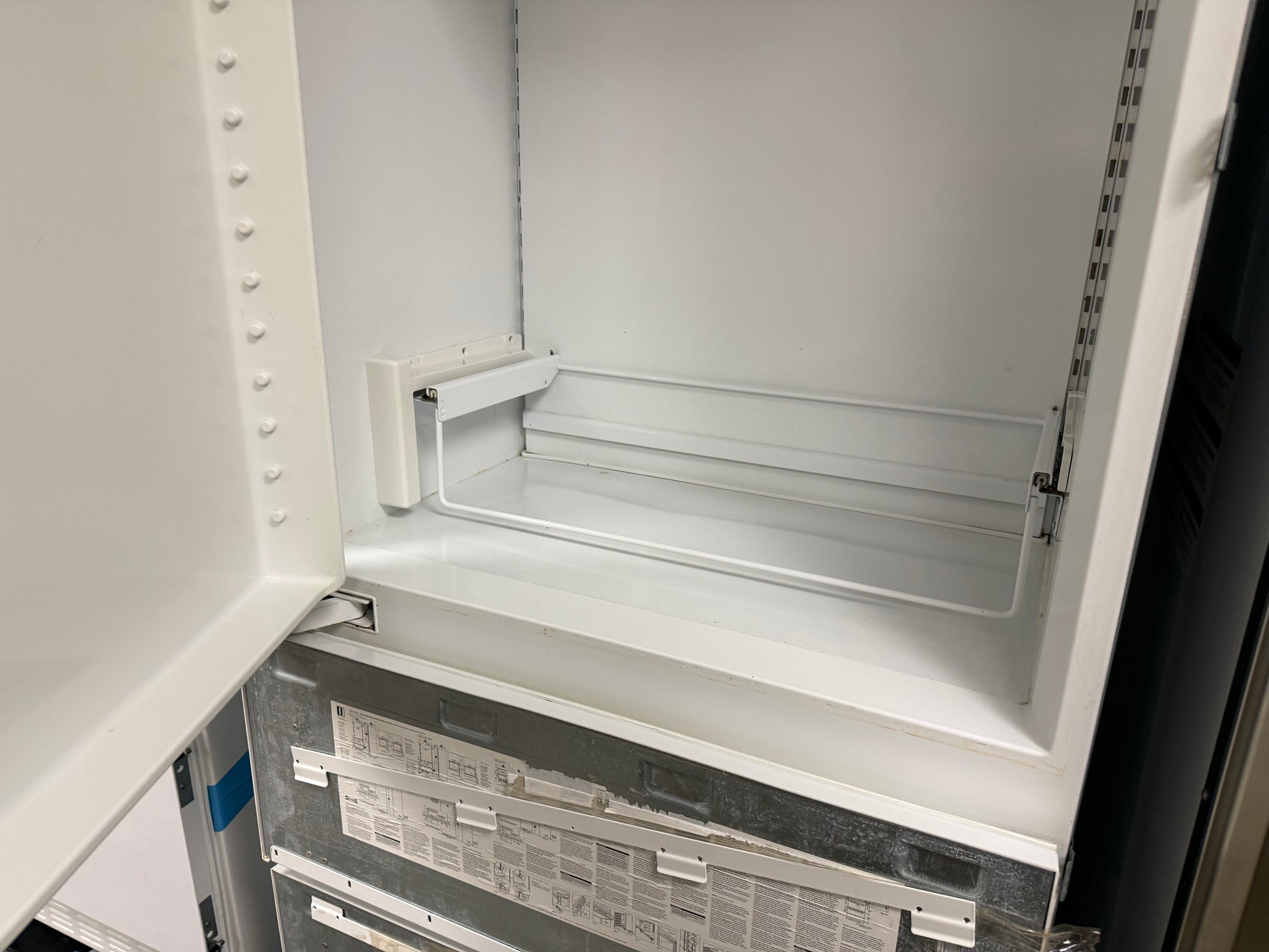 The interior of a white refrigerator or freezer compartment with the door open, showing the empty crisper shelf and metal backing panel.