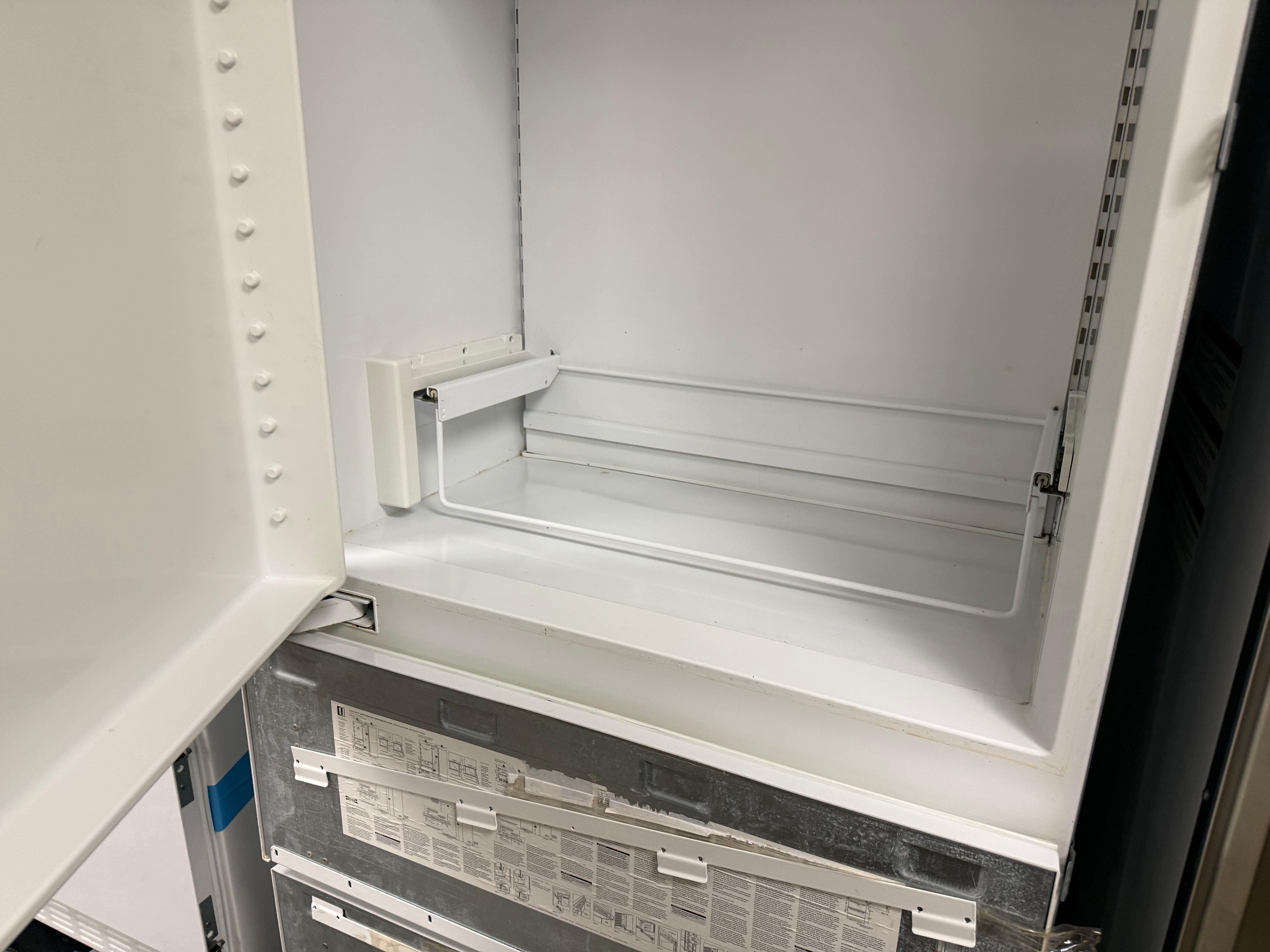 The interior of a white refrigerator or freezer compartment with the door open, showing the empty crisper shelf and metal backing panel.
