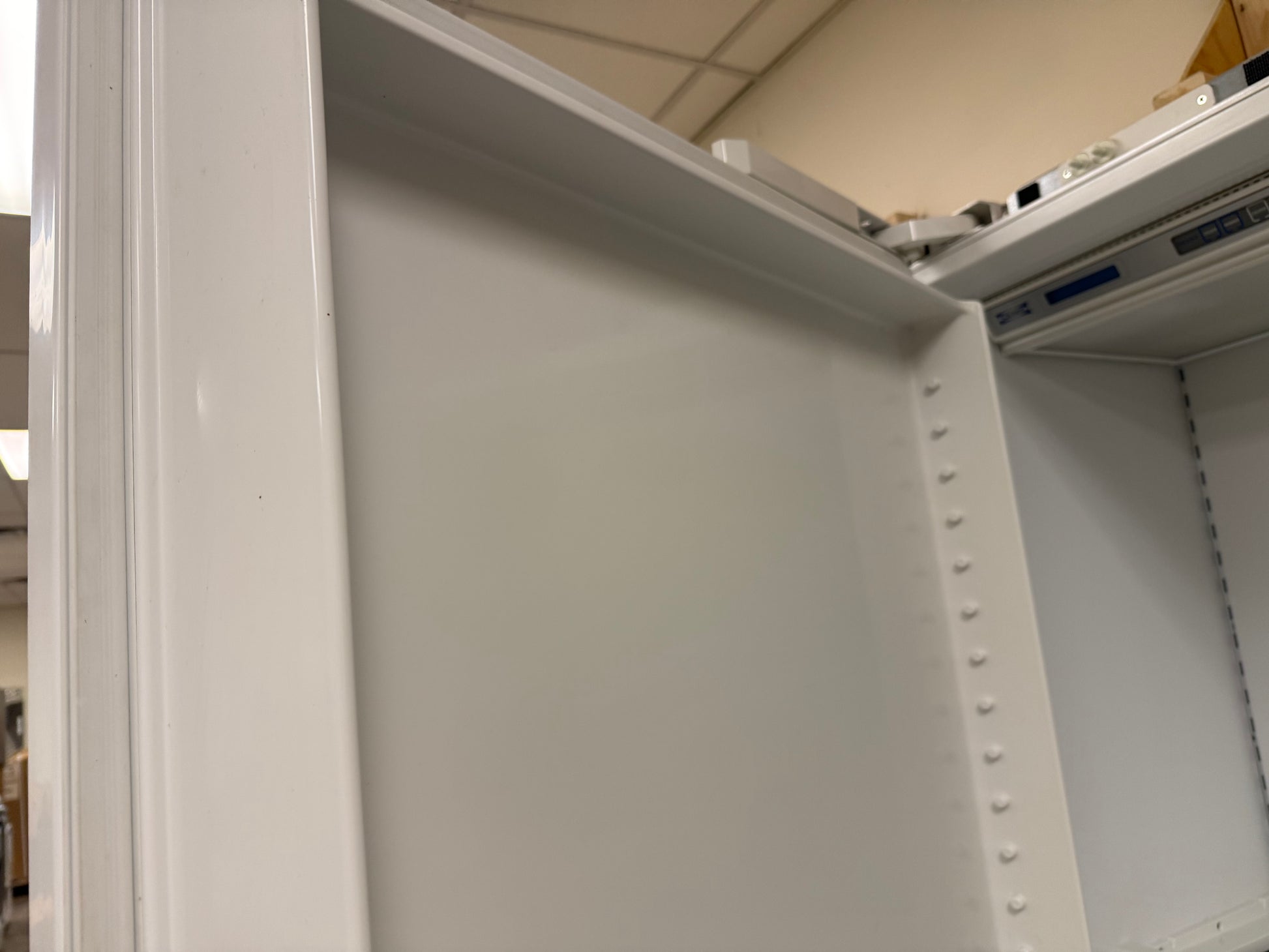 Close-up of the interior corner of a white metal storage cabinet or shelving unit, showing the inner panel, side flange, rivet holes, and part of the top frame in a warehouse setting.