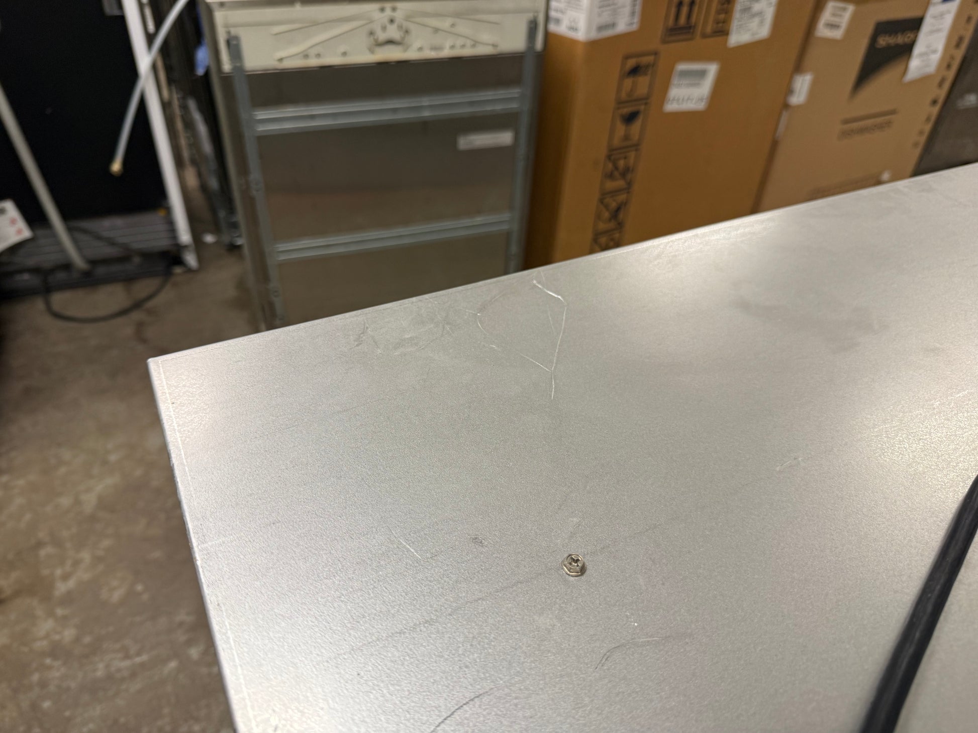 A close-up view of a brushed silver metal surface, likely a workbench or appliance panel, with a small screw visible on top. The background shows a warehouse or storage room setting with cardboard boxes and a metal cart.