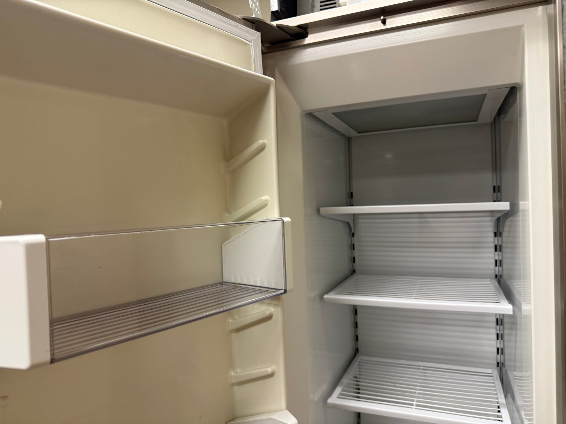 An empty refrigerator interior with cream-colored walls, a clear glass door bin, and white shelves on the main compartment.