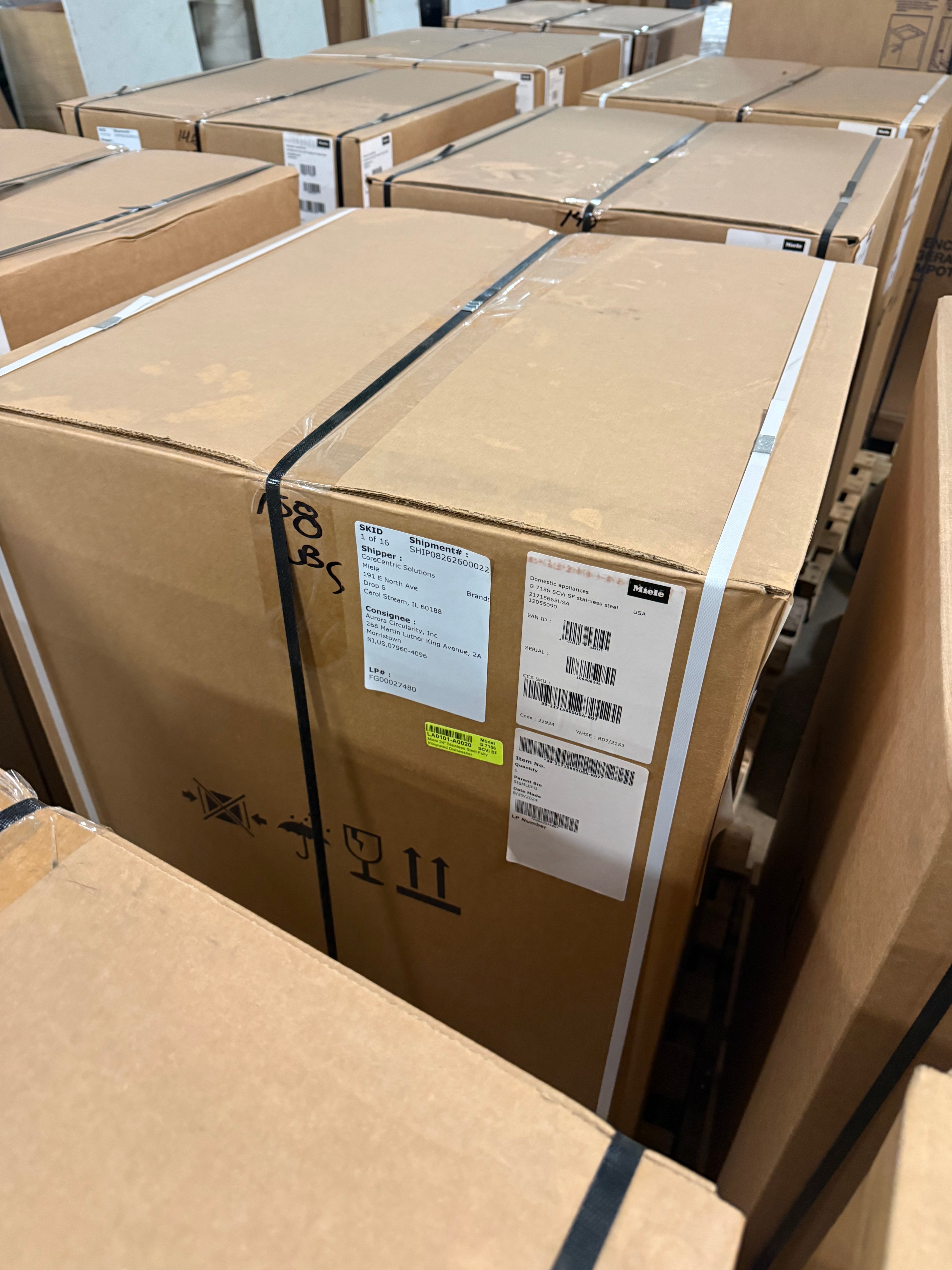 A large brown cardboard shipping box secured with black straps and white tape, with shipping labels attached, photographed in a warehouse among other boxes.