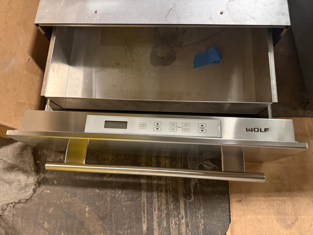 A Wolf commercial-grade stainless steel drawer-style oven or warming drawer with a digital control panel and pull handle, shown partially open on a cardboard box.