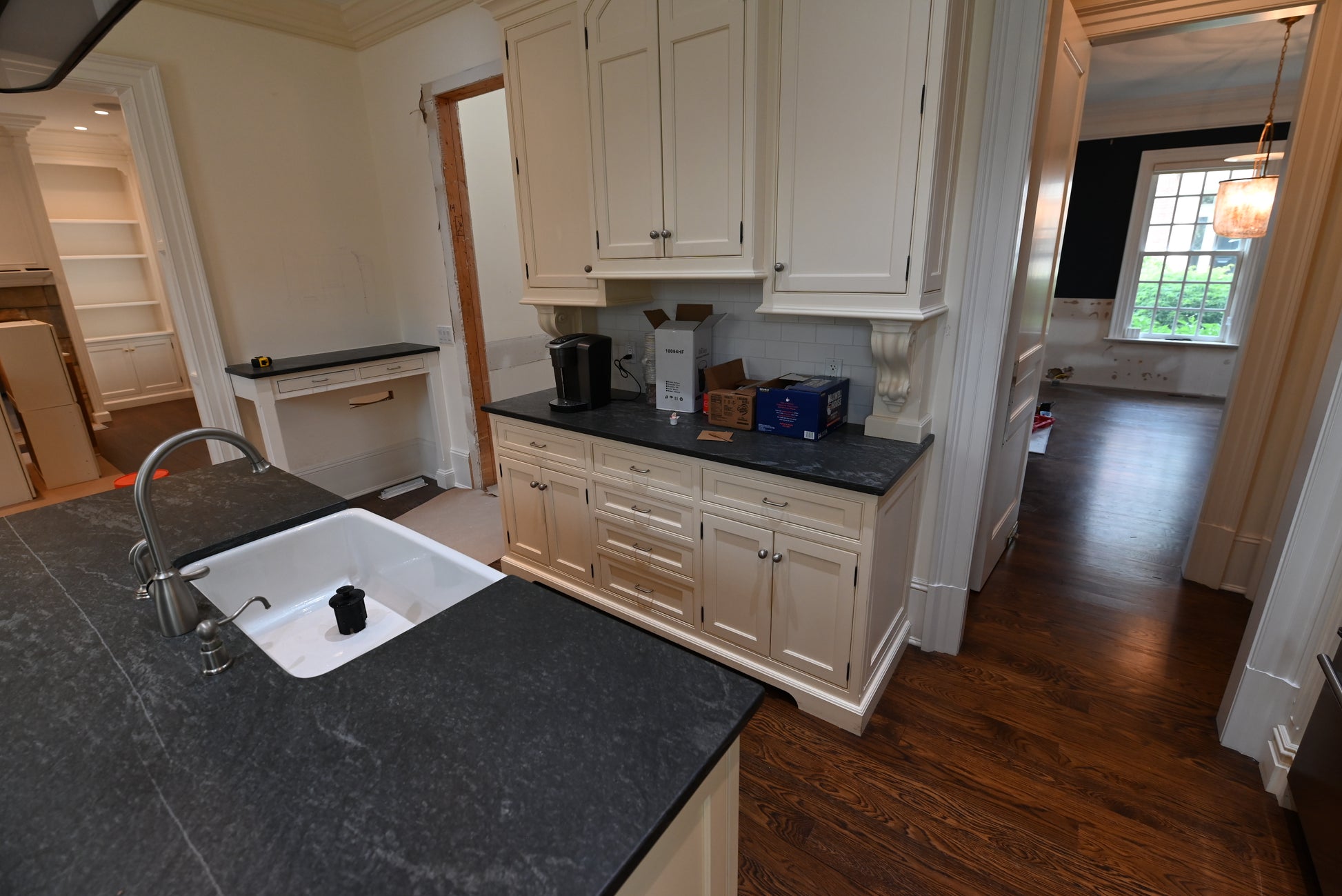 A kitchen interior with cream cabinetry, dark stone countertops, a white sink, and hardwood flooring. A coffee maker and other small appliances sit on the counter.