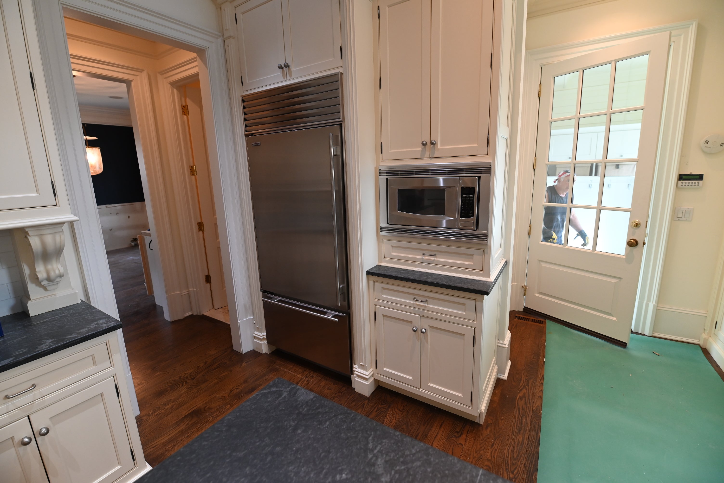 A built-in stainless steel refrigerator and microwave set into white kitchen cabinets with dark countertops, shown in a home interior with hardwood floors and a green carpet runner.