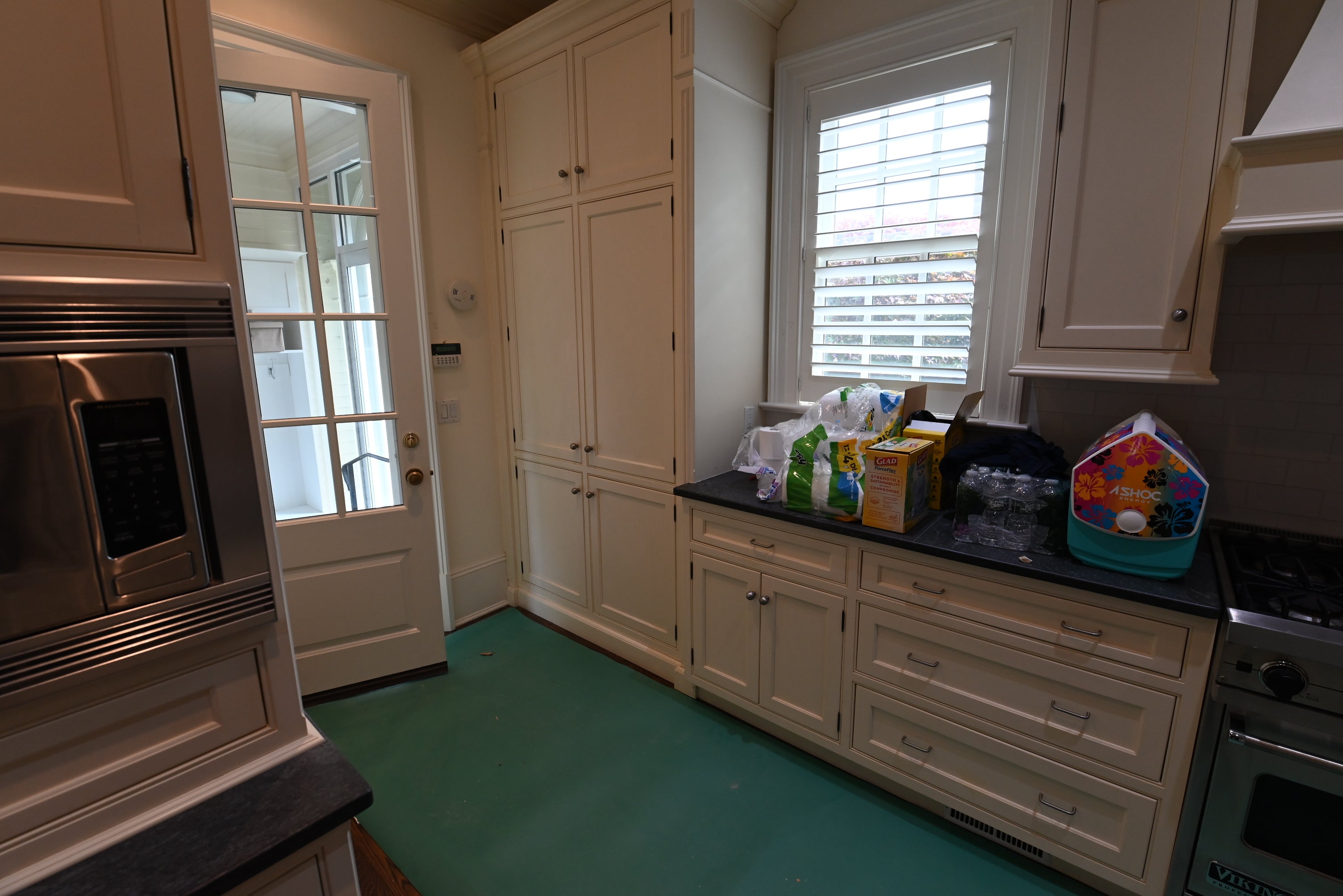 An interior view of a kitchen with cream-colored cabinets, dark countertops, a green floor, a built-in stainless steel microwave on the left, and a window with blinds on the back wall.