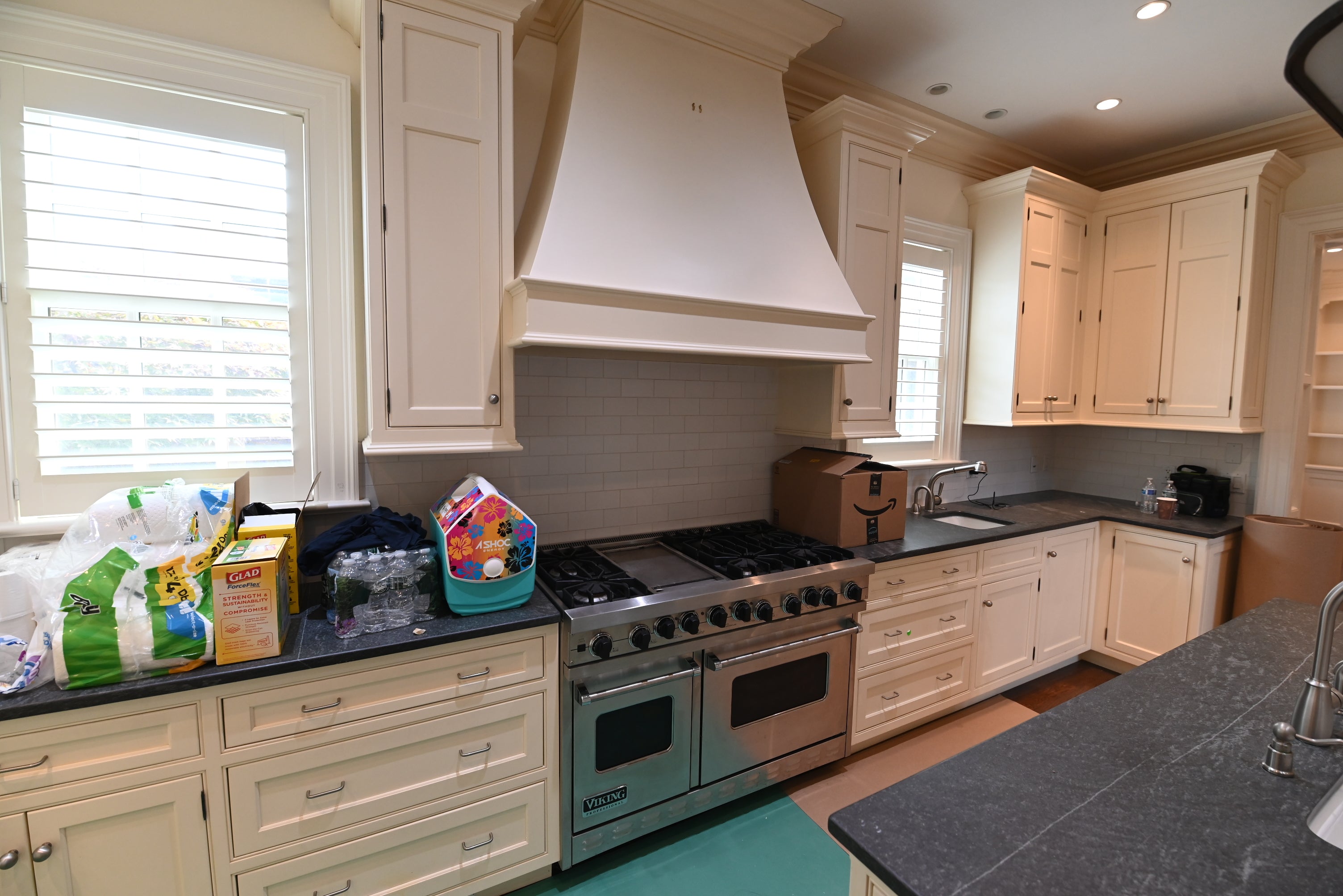 A real kitchen interior with cream cabinets, dark countertops, a stainless steel gas range, a large white range hood, windows with shutters, and a tiled backsplash. No person or animal is present, but this is not a clean isolated product shot; it shows the product in a real environment, so subject_type is Lifestyle.