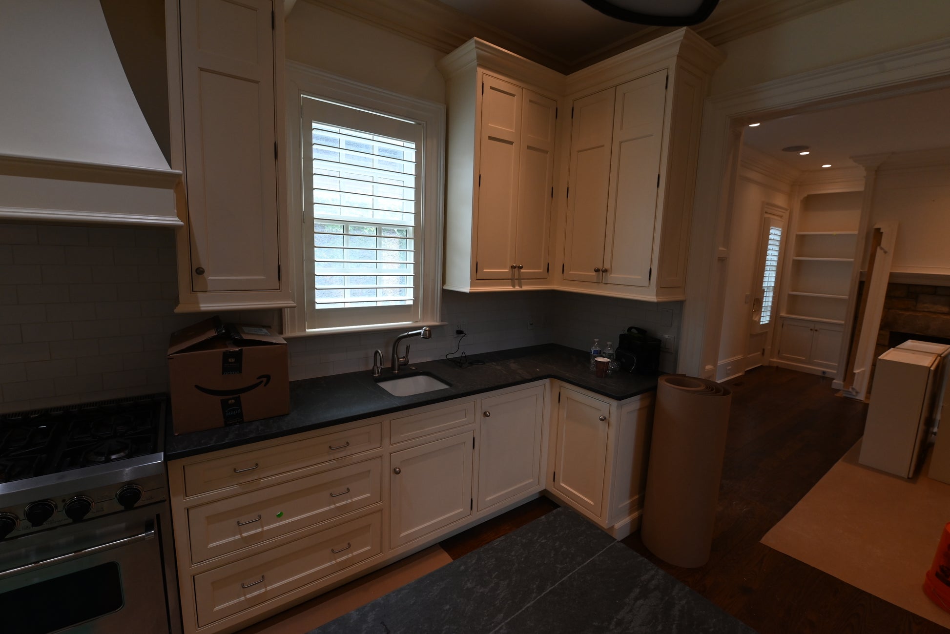 A kitchen interior with cream-colored cabinets, dark countertops, a window with shutters, and a stainless steel stove. A cardboard box with an Amazon smile logo sits on the counter.