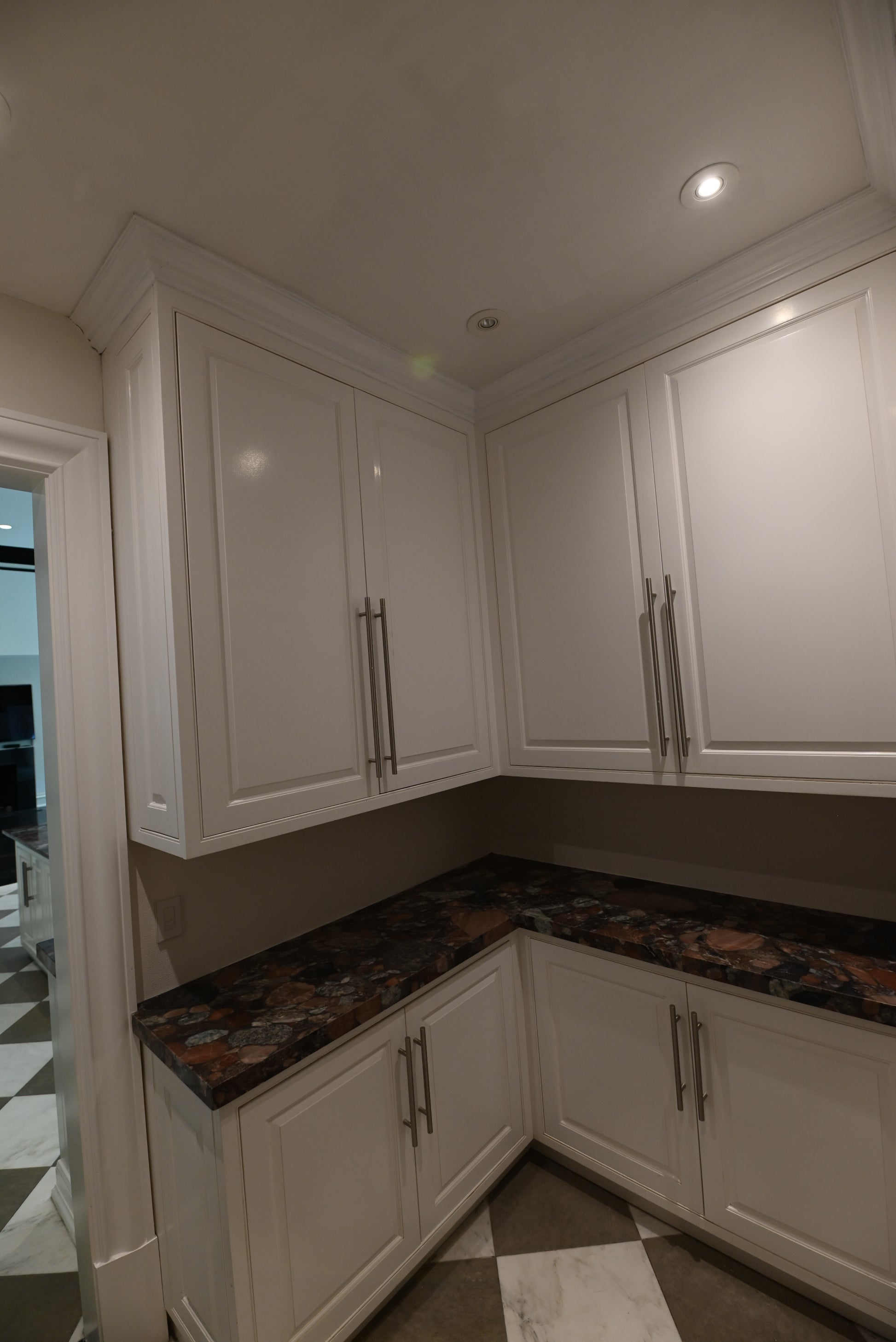 White kitchen cabinets with dark brown and black granite countertops, featuring silver handles and recessed ceiling lights.