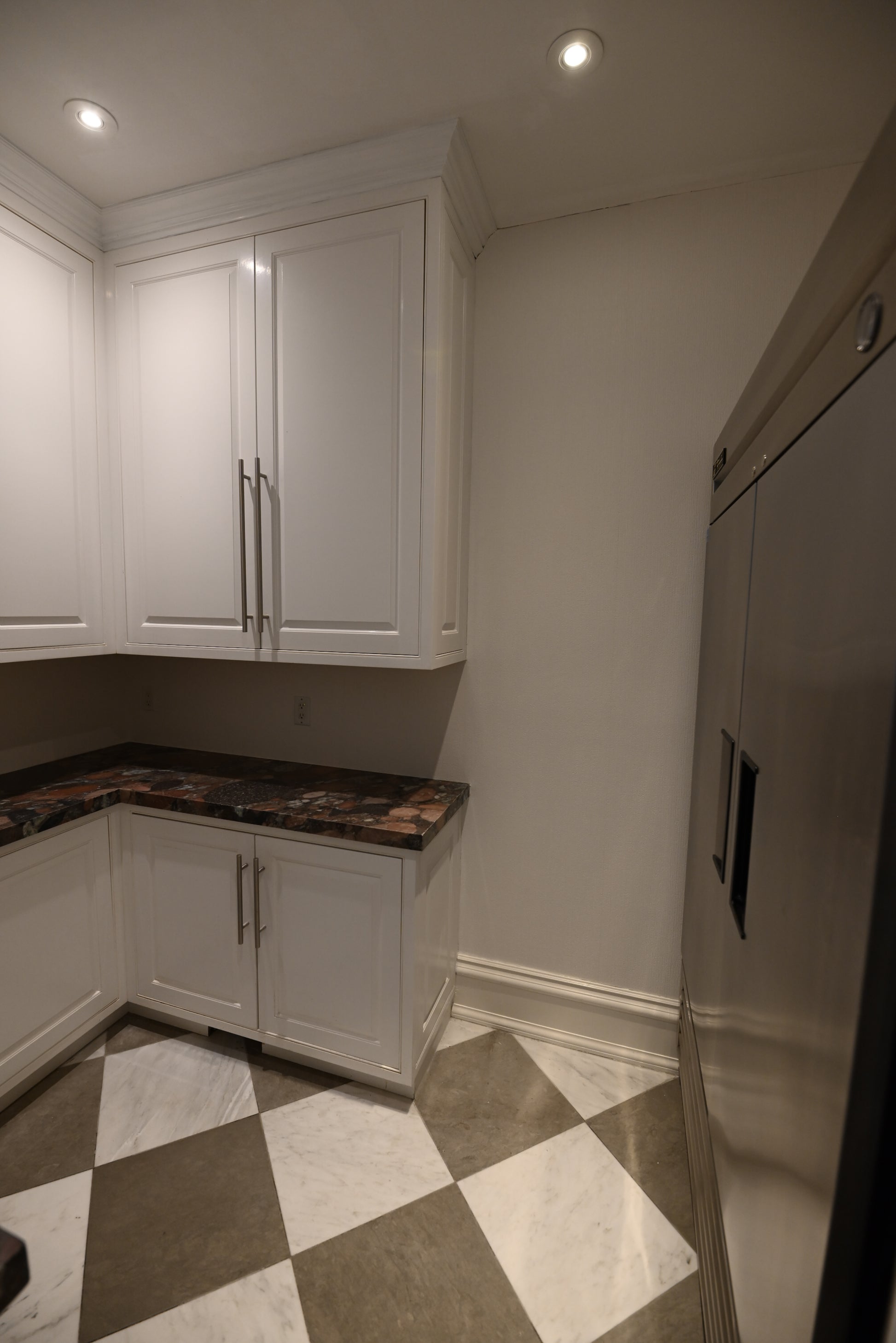 A kitchen corner with white shaker-style cabinets, dark brown marble countertops, and a black-and-white checkered tile floor, next to a stainless steel refrigerator.