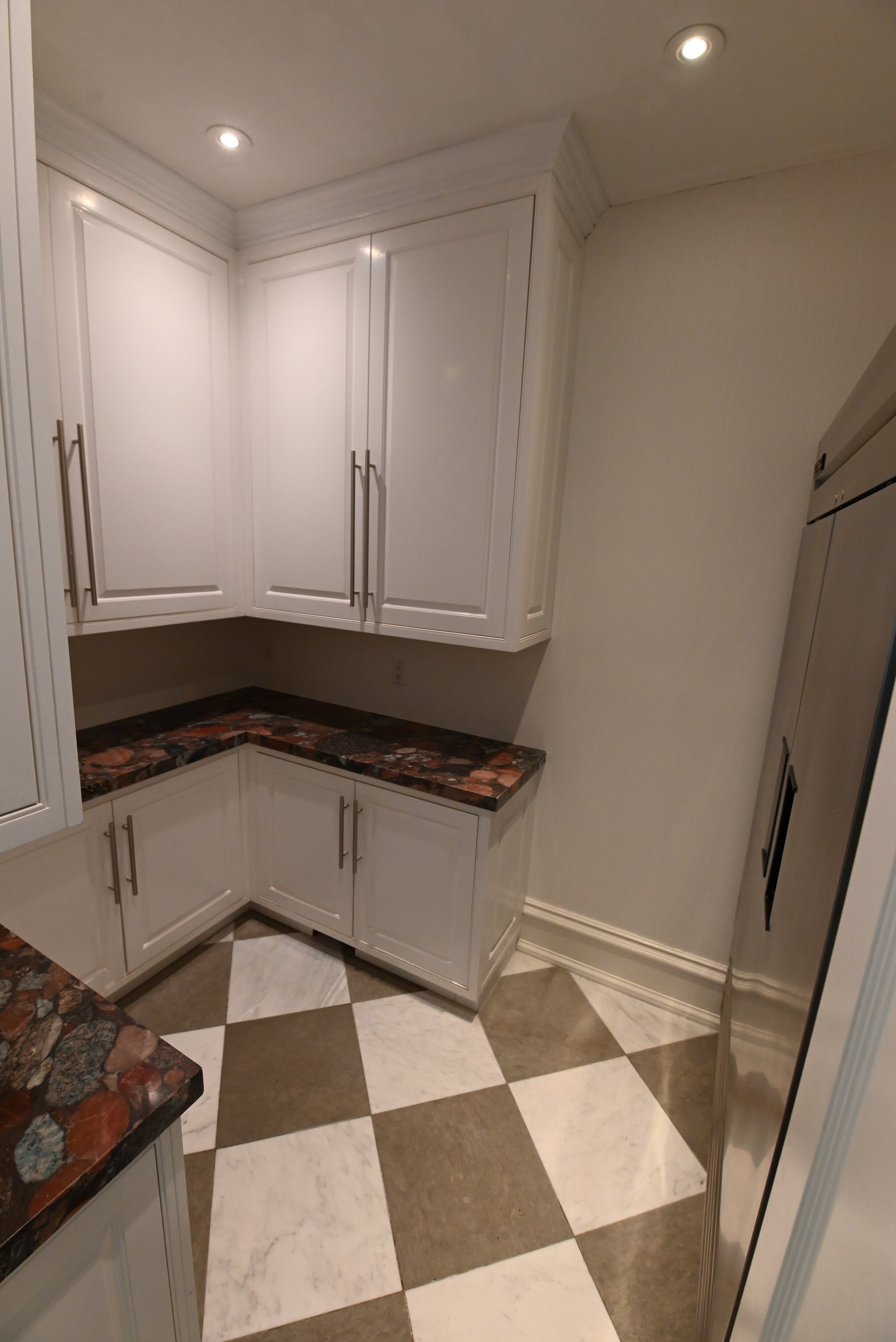 A corner kitchen cabinet setup with white shaker-style cabinets, dark brown and red patterned granite countertops, and a beige and white checkered tile floor.