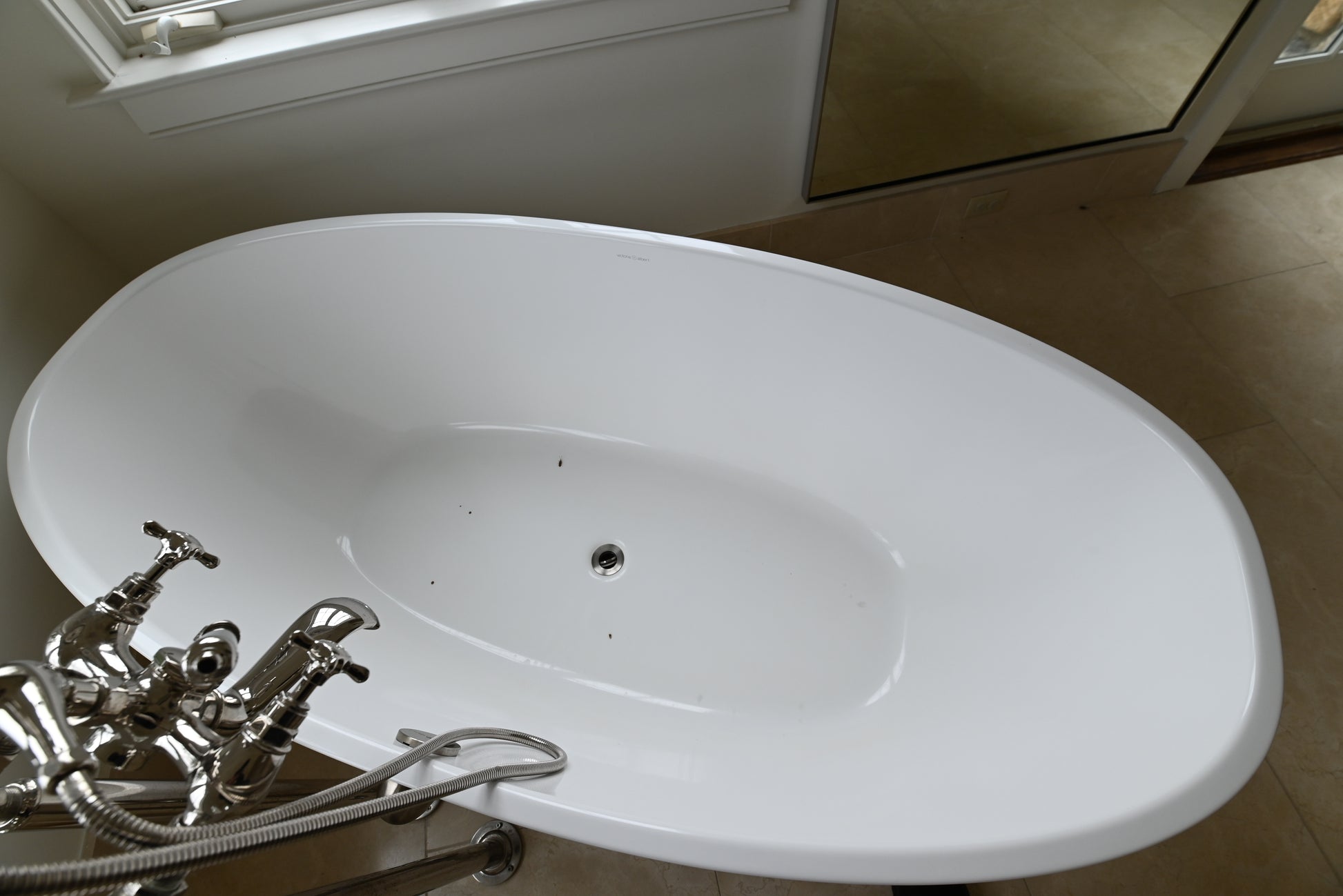 A white freestanding oval bathtub with a polished chrome faucet and hose, photographed in a bathroom setting with tiled floor and a mirror visible.