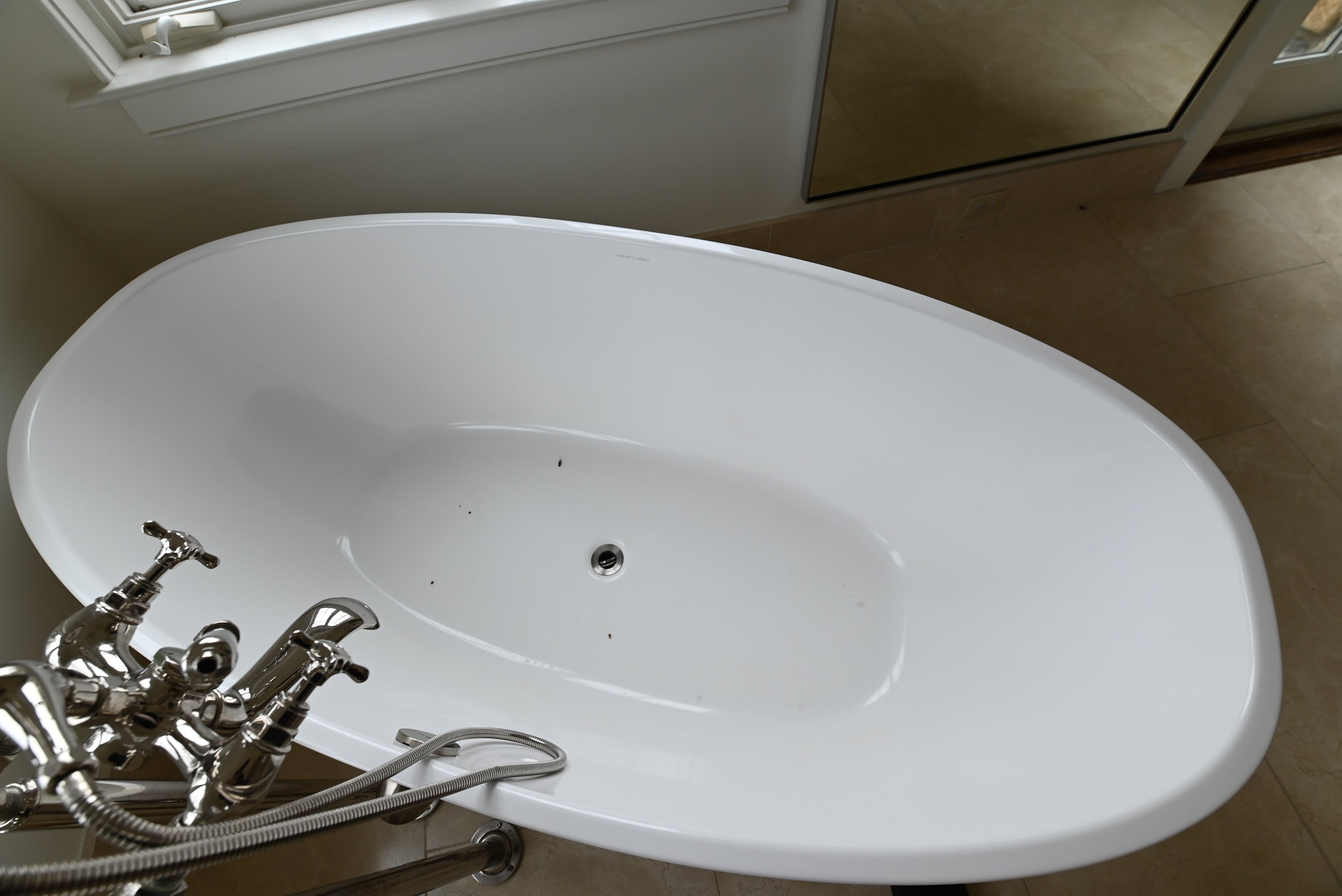 A white freestanding oval bathtub with a polished chrome faucet and hose, photographed in a bathroom setting with tiled floor and a mirror visible.