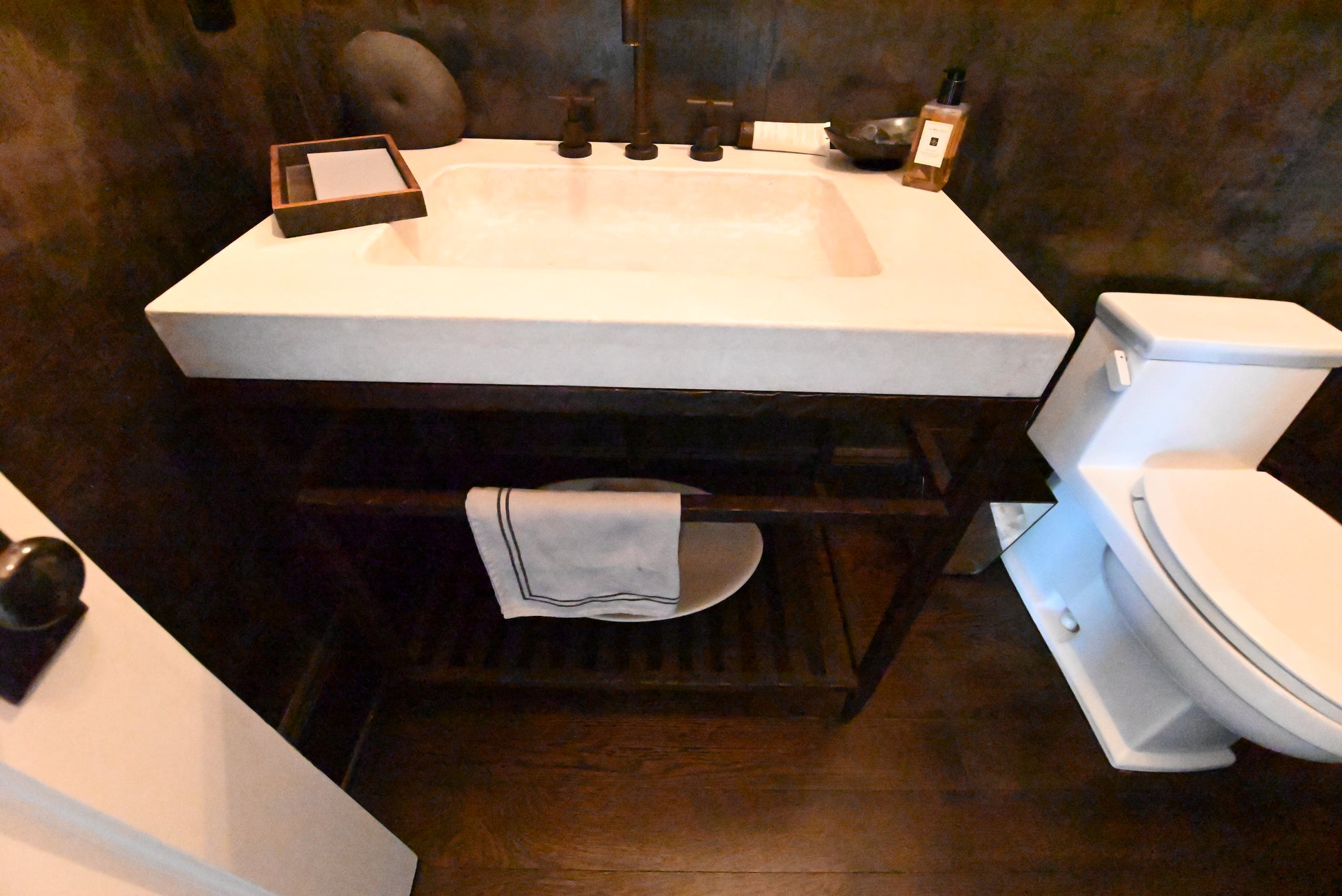 A modern bathroom vanity with a white rectangular stone sink and dark wood open shelving, shown next to a white toilet in a bathroom with dark wood floors and textured walls.