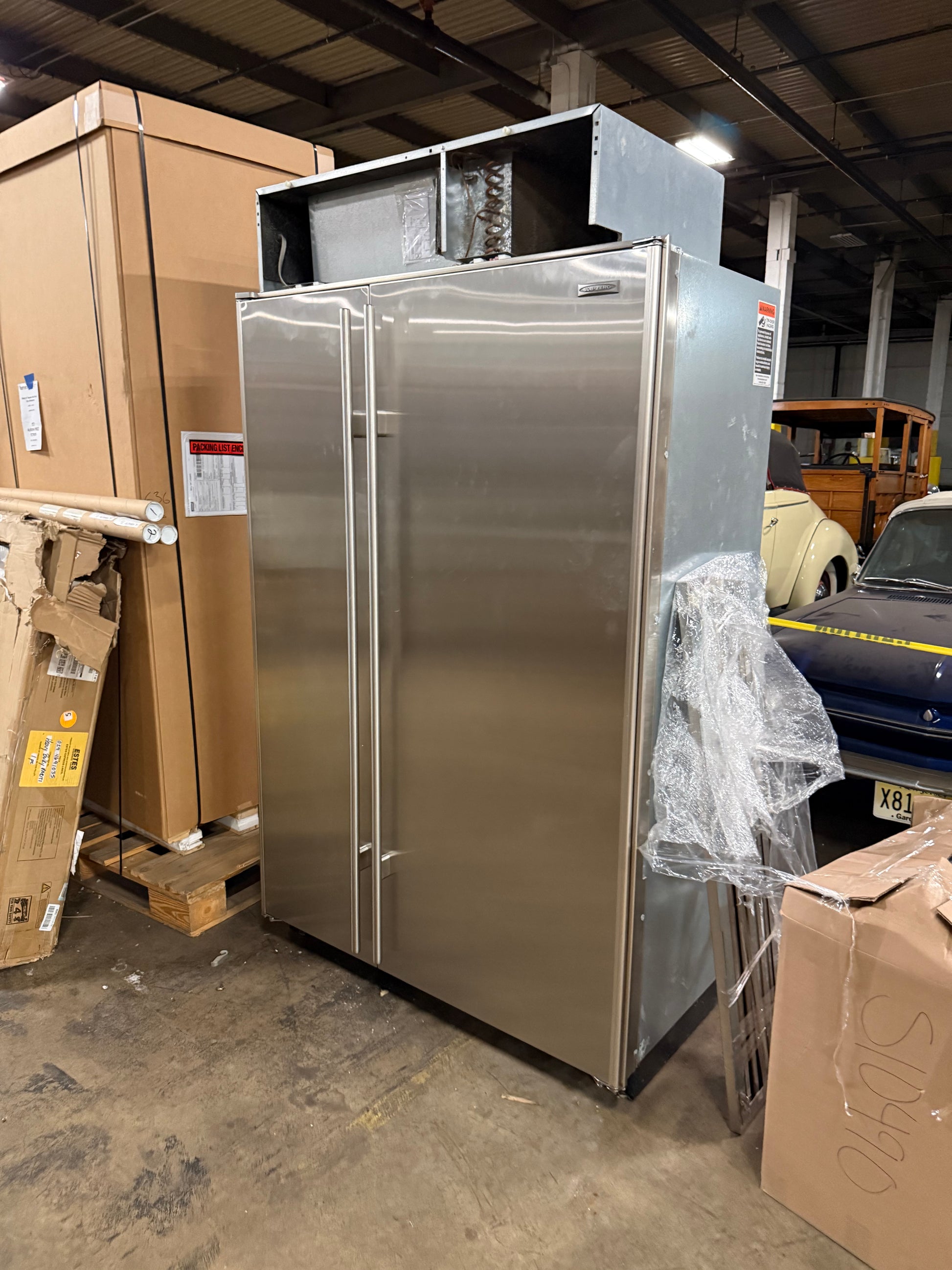 A stainless steel commercial refrigerator or freezer unit with two front drawers, photographed in a warehouse setting among cardboard boxes.
